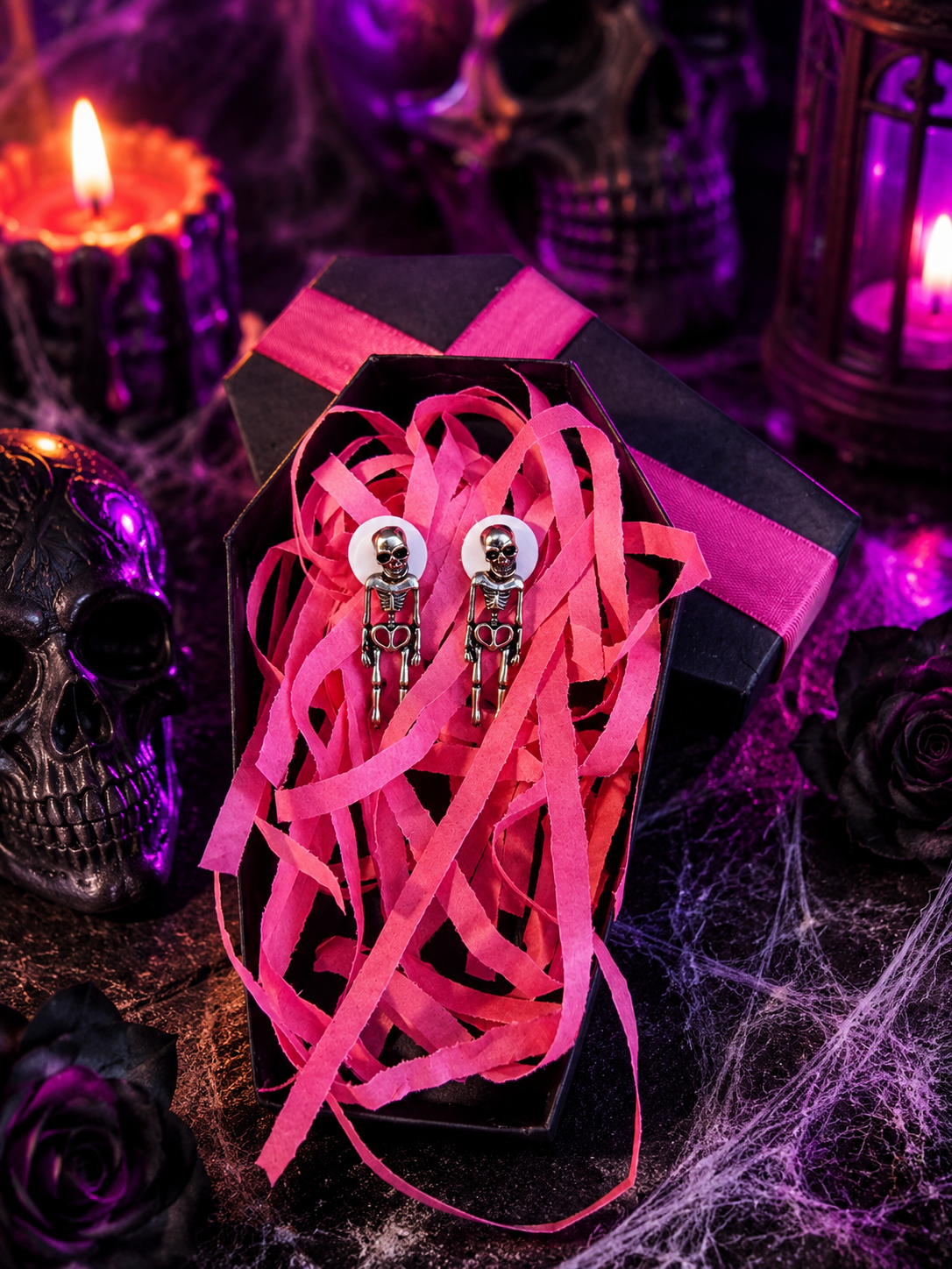 A pair of gothic skeleton stud earrings displayed in an open black coffin-shaped box filled with bright pink ribbon, surrounded by Halloween-themed decor including a skull, candles, roses, and cobwebs.