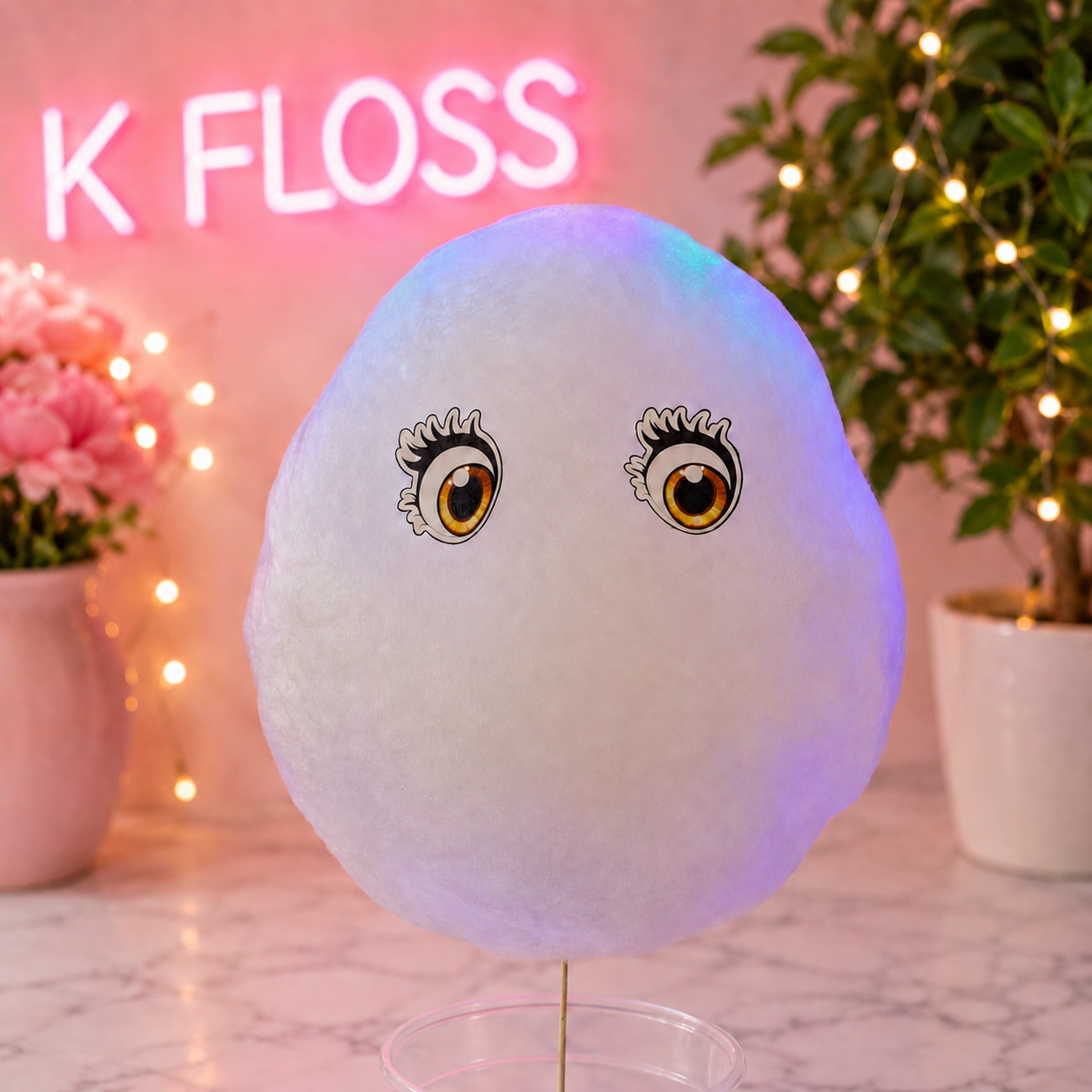 A fluffy white cotton candy ball with printed cartoon eyes sits on a clear stand on a marble surface, with pink neon text and potted plants in the background.