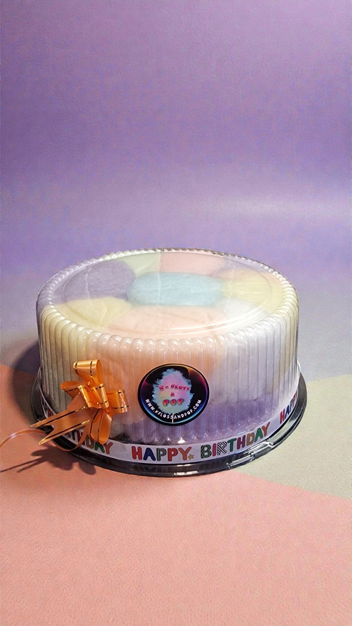 Round cake with colorful layers in a clear container on a pink surface with a purple background