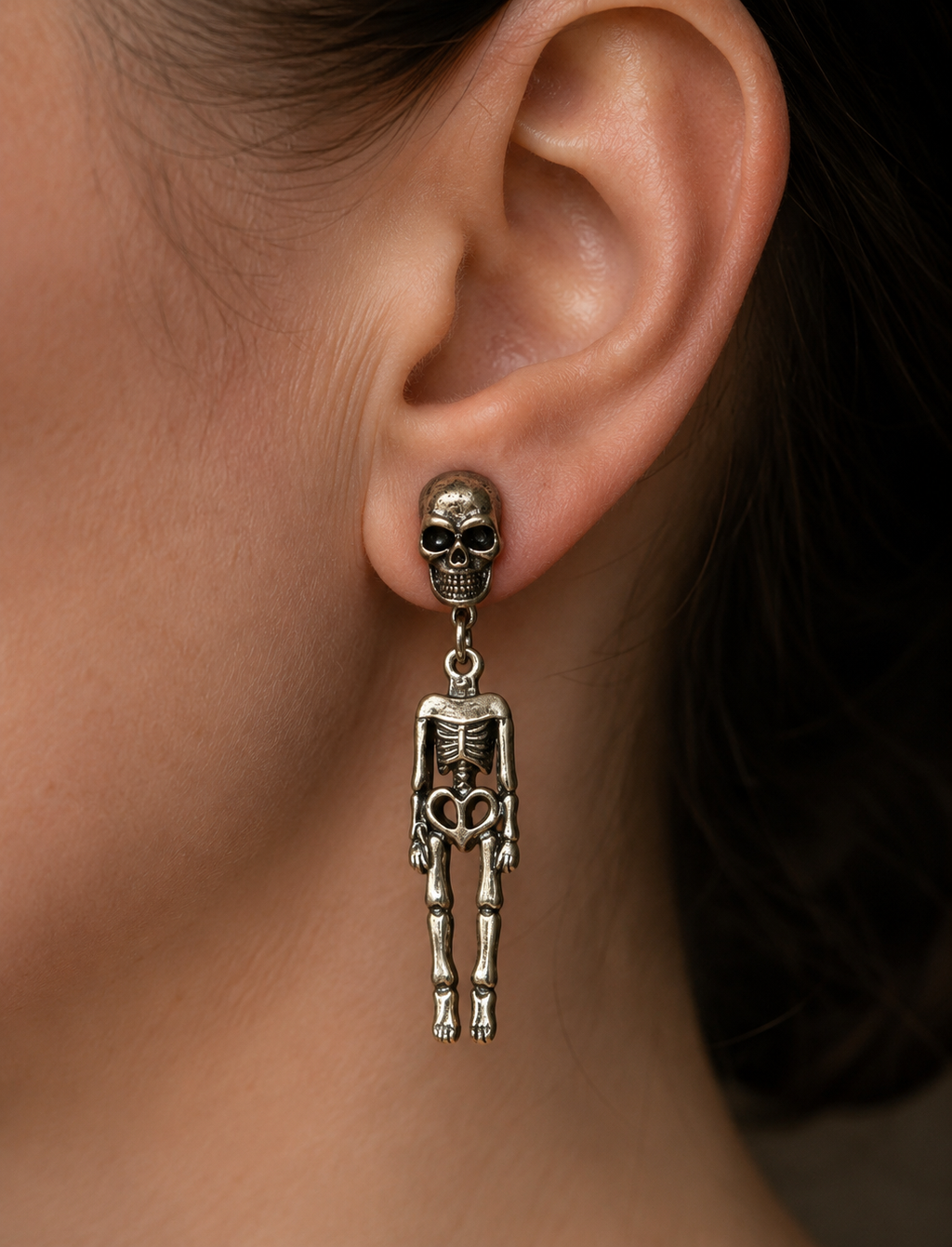 A close-up of a person's ear wearing a dangling skeleton earring. The earring features a small skull at the top with a dangling skeleton figure below, in a silver-tone metal finish.