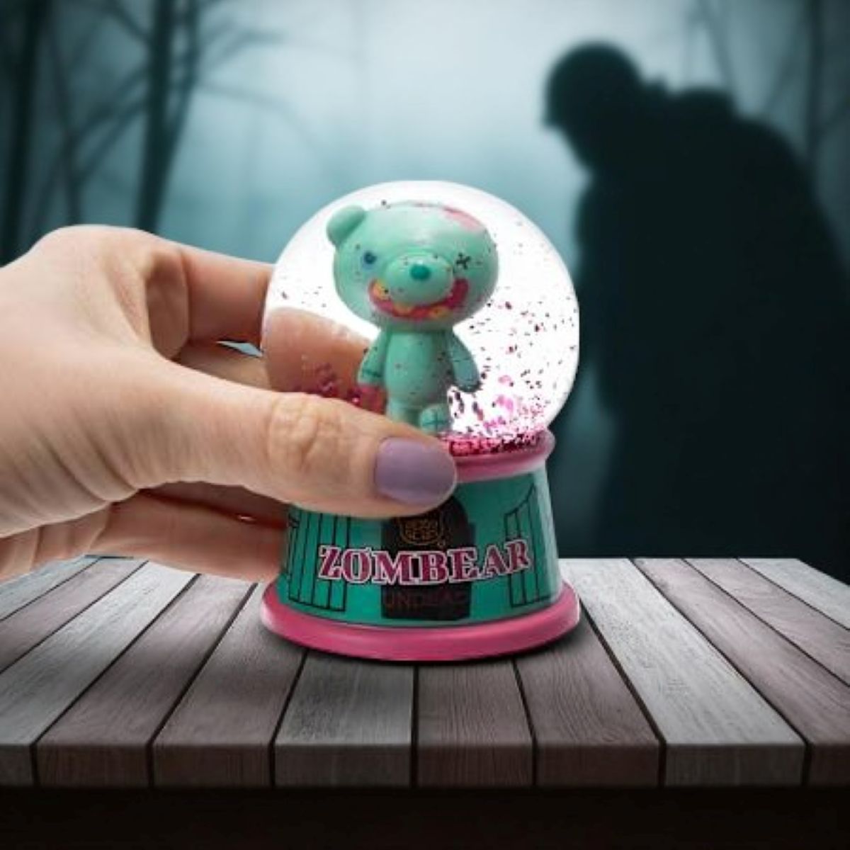 Alt text: Hand holding Deddy Bears Zombiebear Mini Snow Globe with green bear inside, pink and teal base, spooky background.