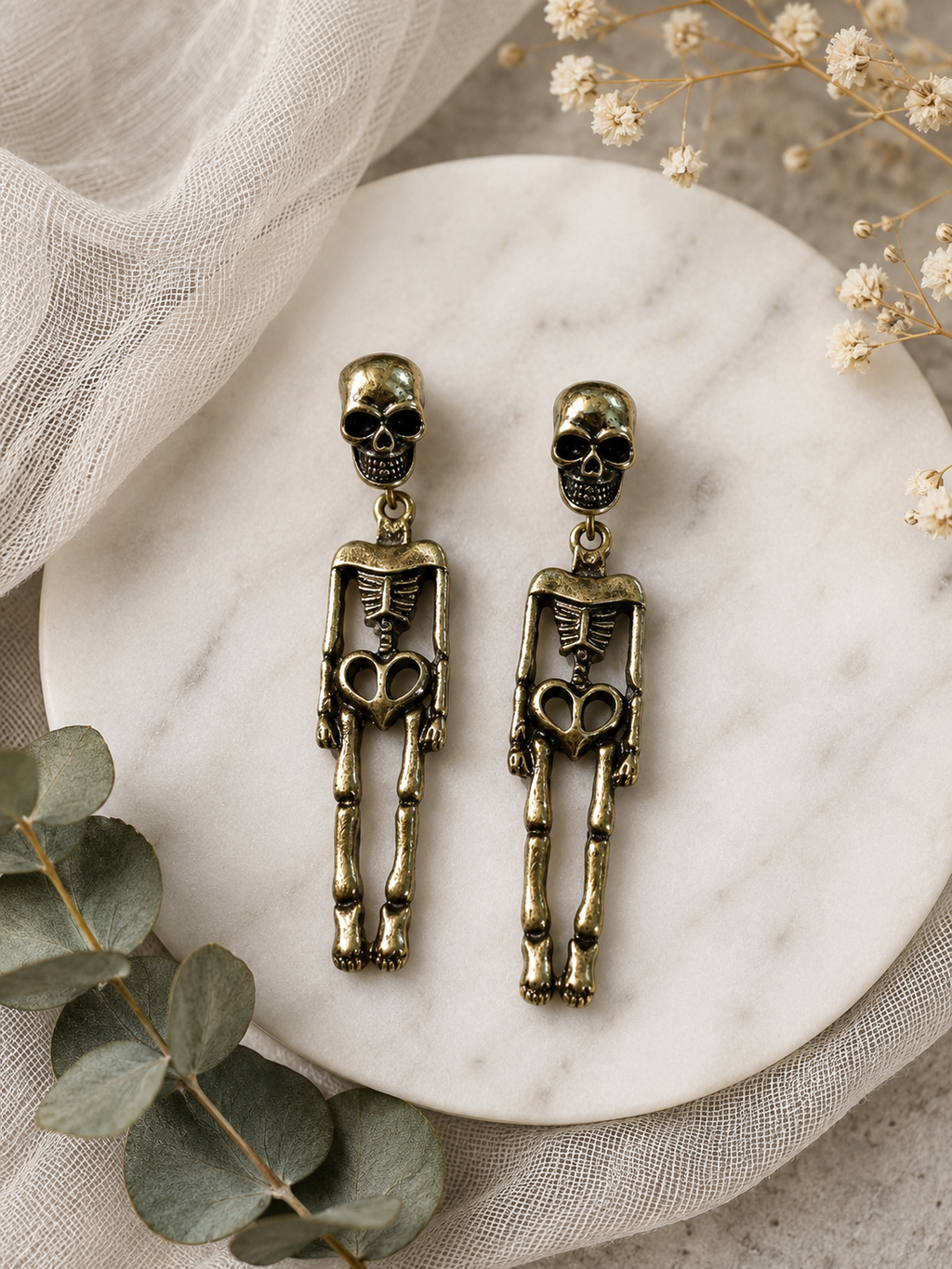 A pair of dangling skeleton earrings in an antique bronze finish, displayed on a round white marble plate with eucalyptus leaves and dried flowers.