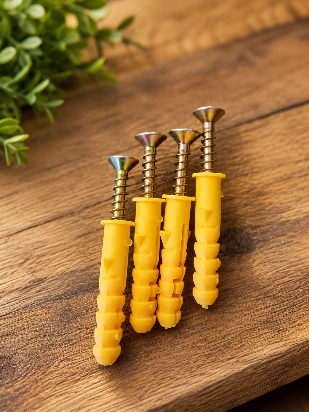 Four yellow wall anchors with metallic screws arranged on a rustic wooden surface with a blurred green plant in the background.