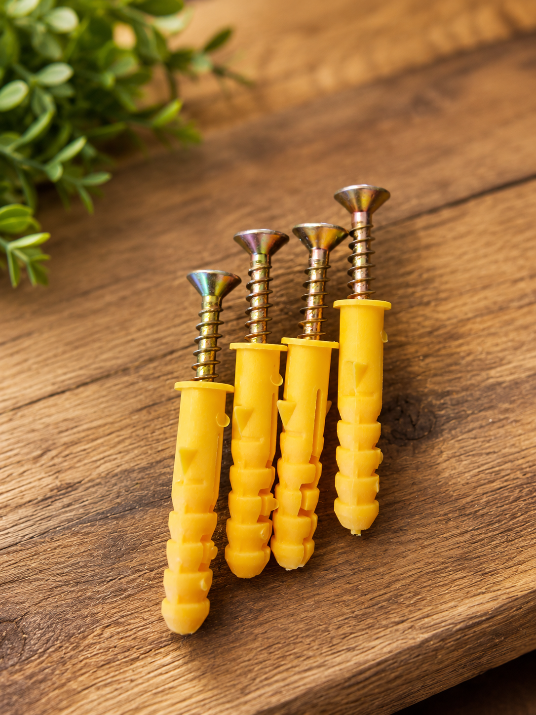 Four yellow wall anchors with metallic screws arranged on a rustic wooden surface with a blurred green plant in the background.