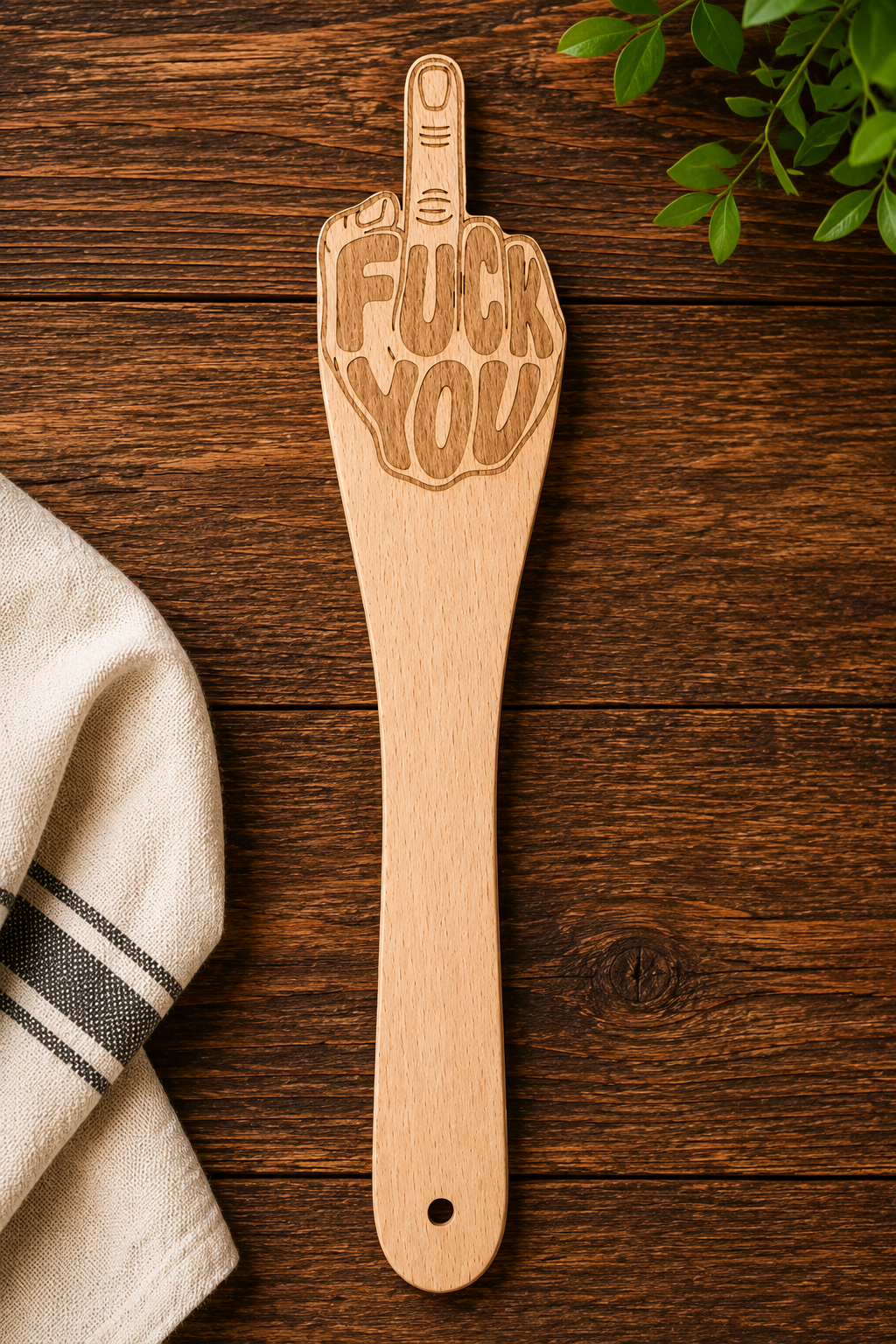 A wooden kitchen utensil shaped like a hand gesturing with the middle finger, engraved with the words 'FUCK YOU', displayed on a dark wood background next to a folded kitchen towel and green leaves.