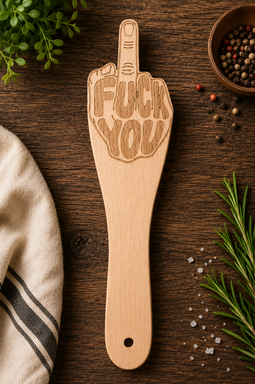 A wooden spatula-shaped cutting board with a hand gesture and the words 'FUCK YOU' engraved on it, displayed on a dark wooden tabletop with herbs, salt, peppercorns, and a striped cloth.
