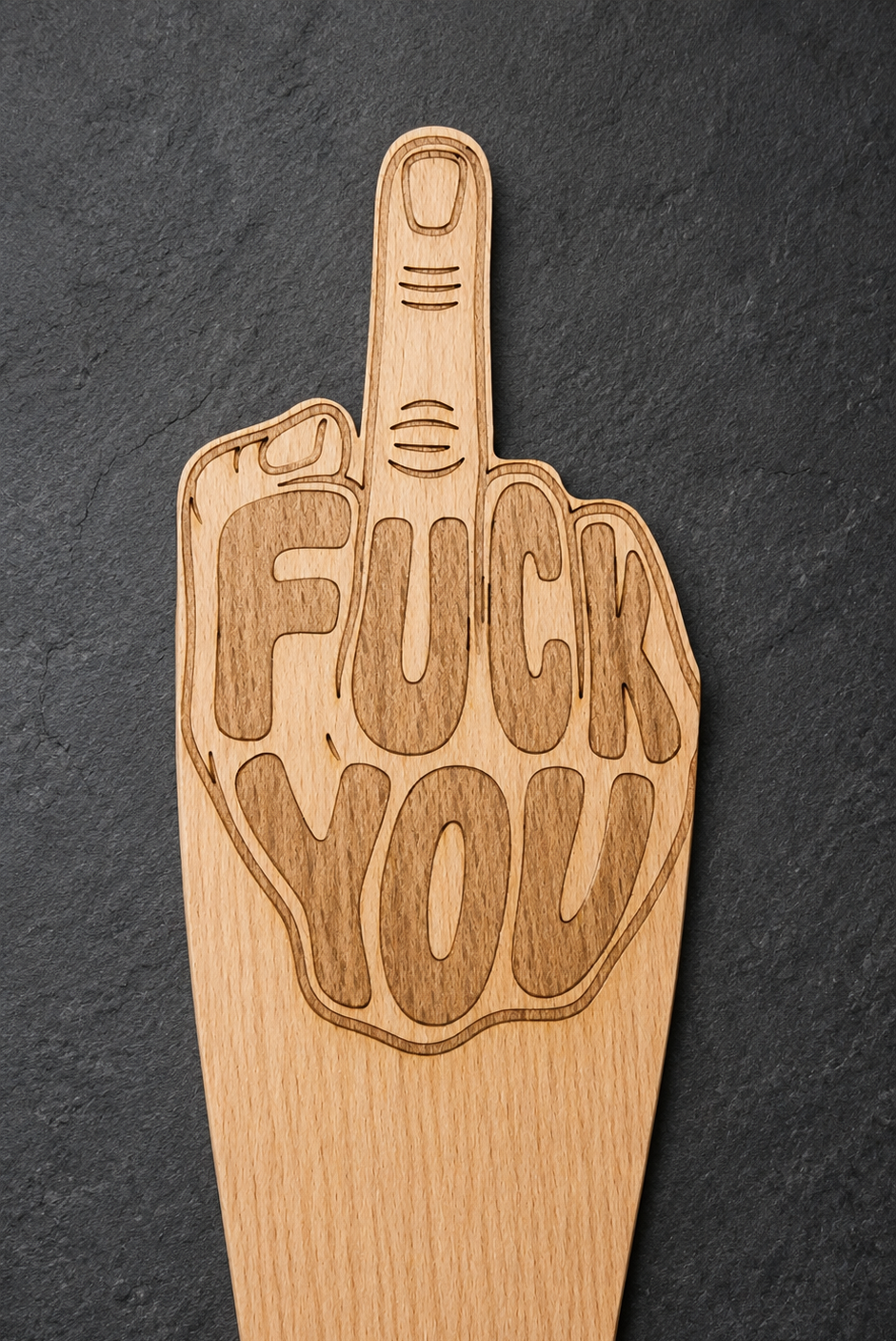 A wooden spatula shaped like a hand giving the middle finger, with the words 'FUCK YOU' engraved on the palm, shown on a dark slate background.