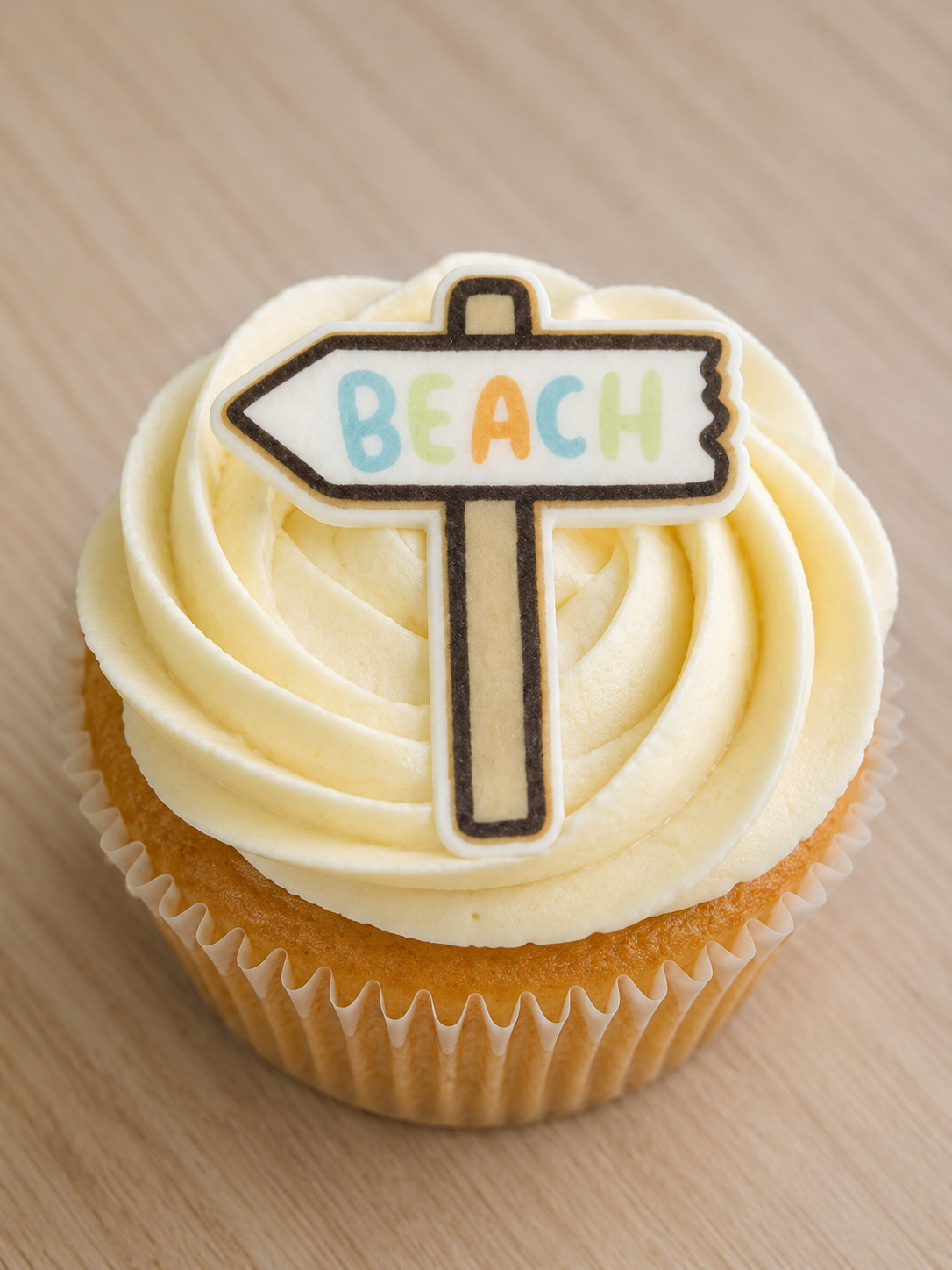 A frosted cupcake with pale cream swirls and a small sign-shaped topper that reads “BEACH” in multicolored letters, set on a light wood surface.