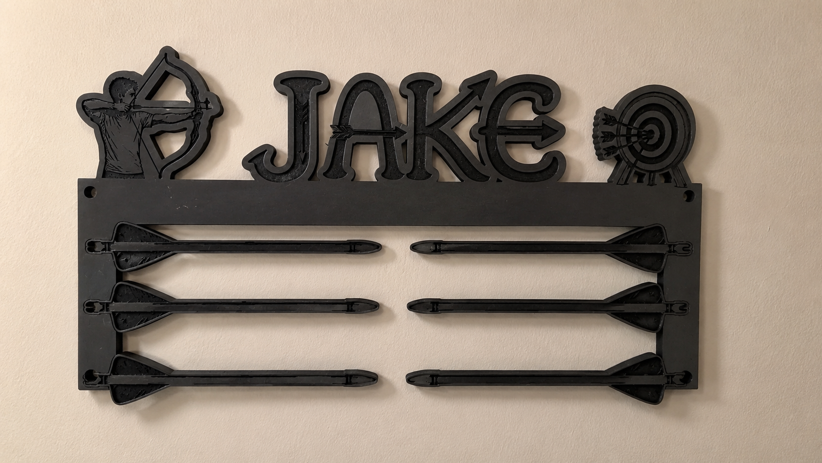 A black metal wall-mounted arrow rack with the name 'JAKE' in raised letters, flanked by a bow and arrow motif on the left and a target symbol on the right, holding six arrows against a beige wall.