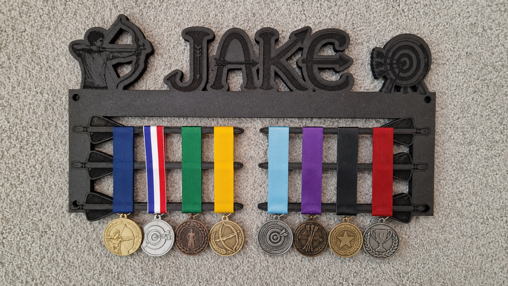 A black metal wall-mounted display rack with the name 'JAKE' across the top and multiple ribbon medals hanging below, photographed against a light textured wall.
