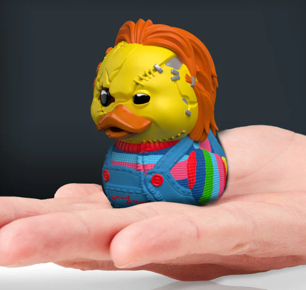 Yellow rubber duck with orange hair and colorful outfit held in a hand against a dark background