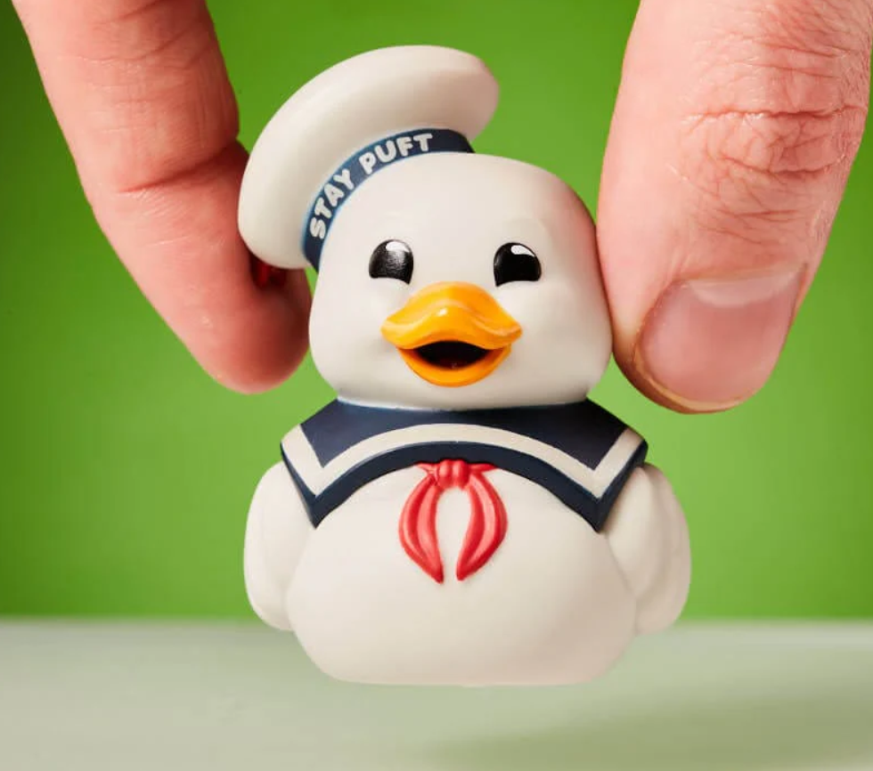 Mini Stay Puft TUBBZ duck collectible toy dressed as the Ghostbusters marshmallow sailor, held between two fingers on a green background.