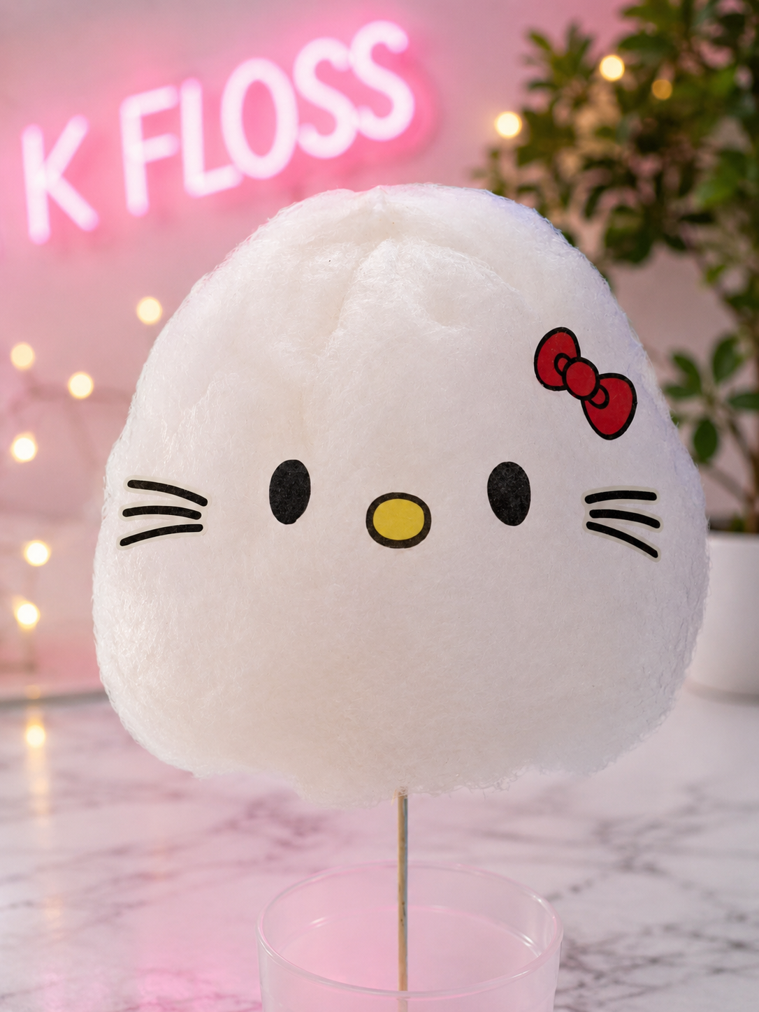 A white Hello Kitty cotton candy ball on a stick, displayed on a pink stand with a pink Hello Kitty sign and fairy lights in the background.