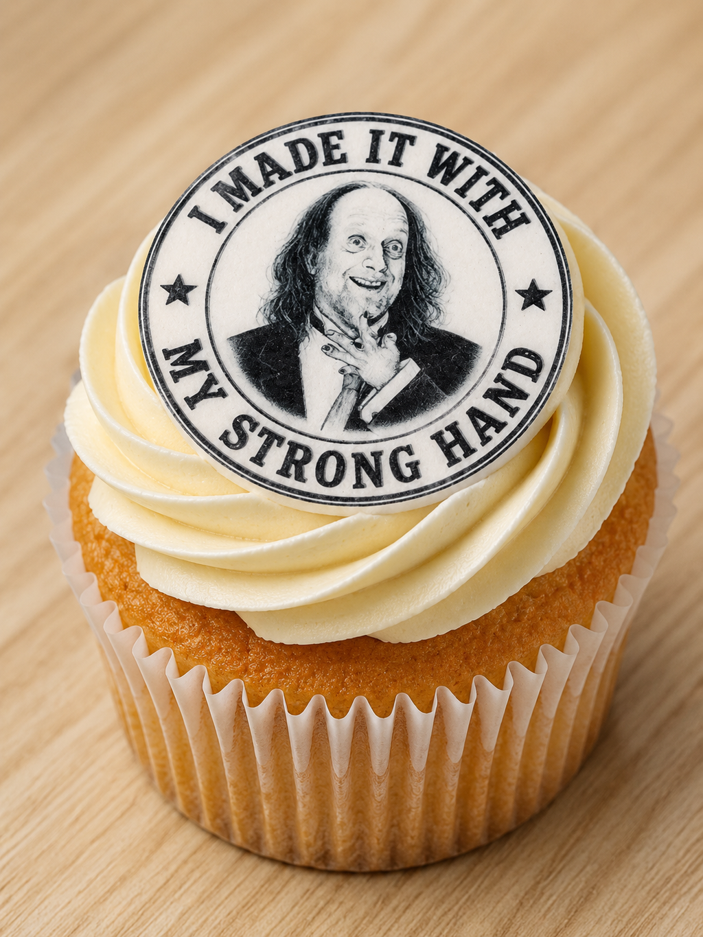 A close-up of a frosted cupcake topped with a round black-and-white printed topper that reads “I MADE IT WITH MY STRONG HAND” and shows a portrait. The cupcake sits on a light wood surface.