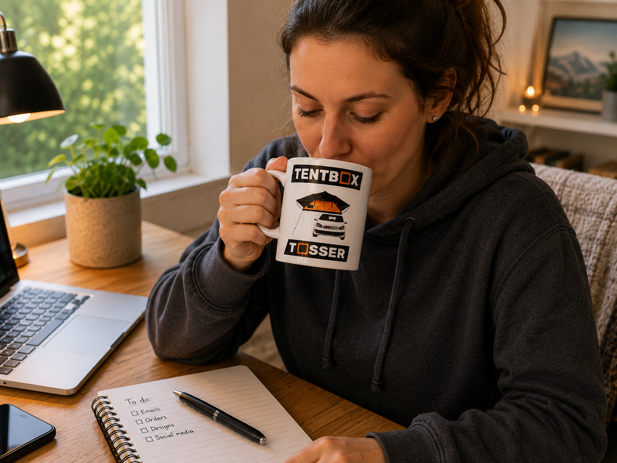 A woman in a dark gray hoodie sits at a wooden desk, drinking from a white mug printed with the brand name TENTBOX and a tent graphic. The scene is a cozy home office with a laptop, notebook, and plant in the background.