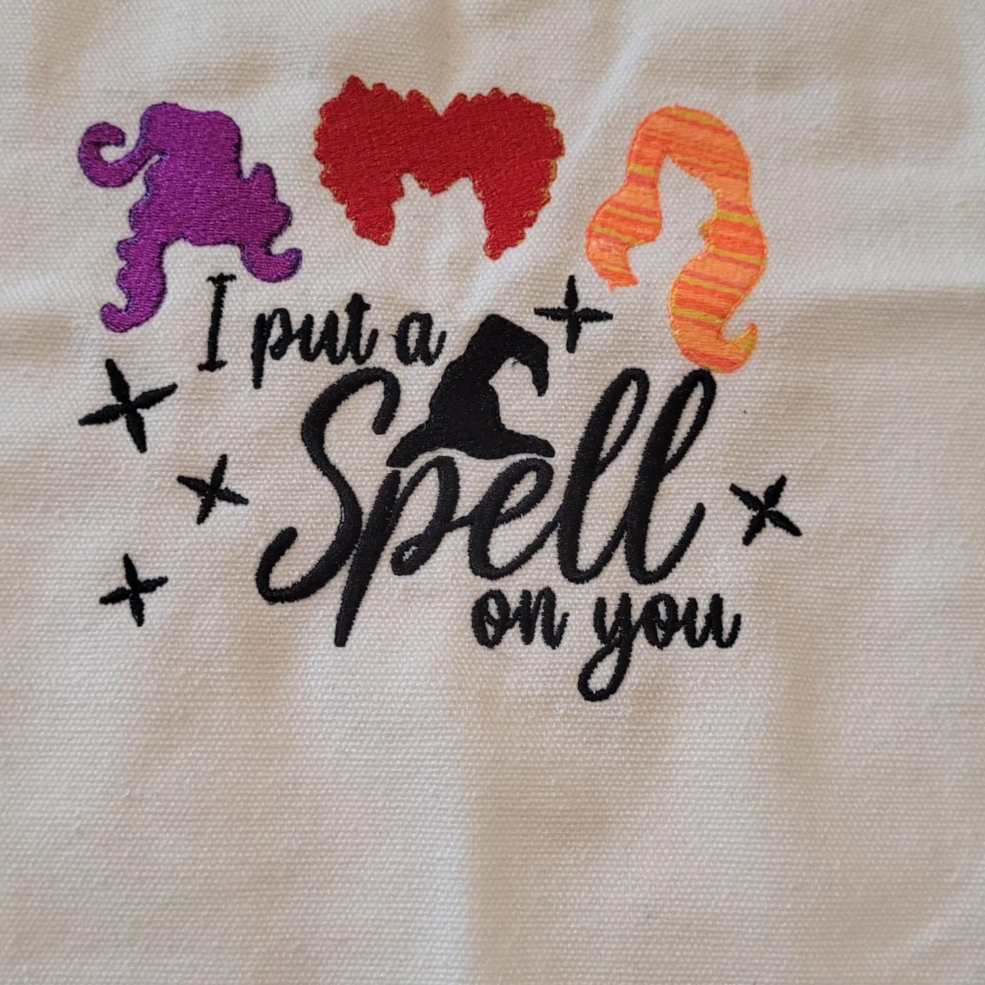 Embroidered design with colorful hair styles and text 'I put a spell on you' on a beige fabric background.