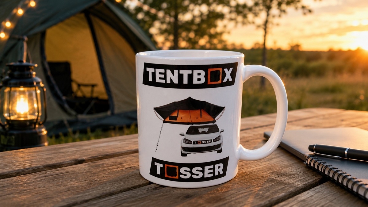 A white ceramic mug with a black and orange tentbox graphic sits on a wooden table outdoors at sunset, with a tent, lantern, and notebook in the background.