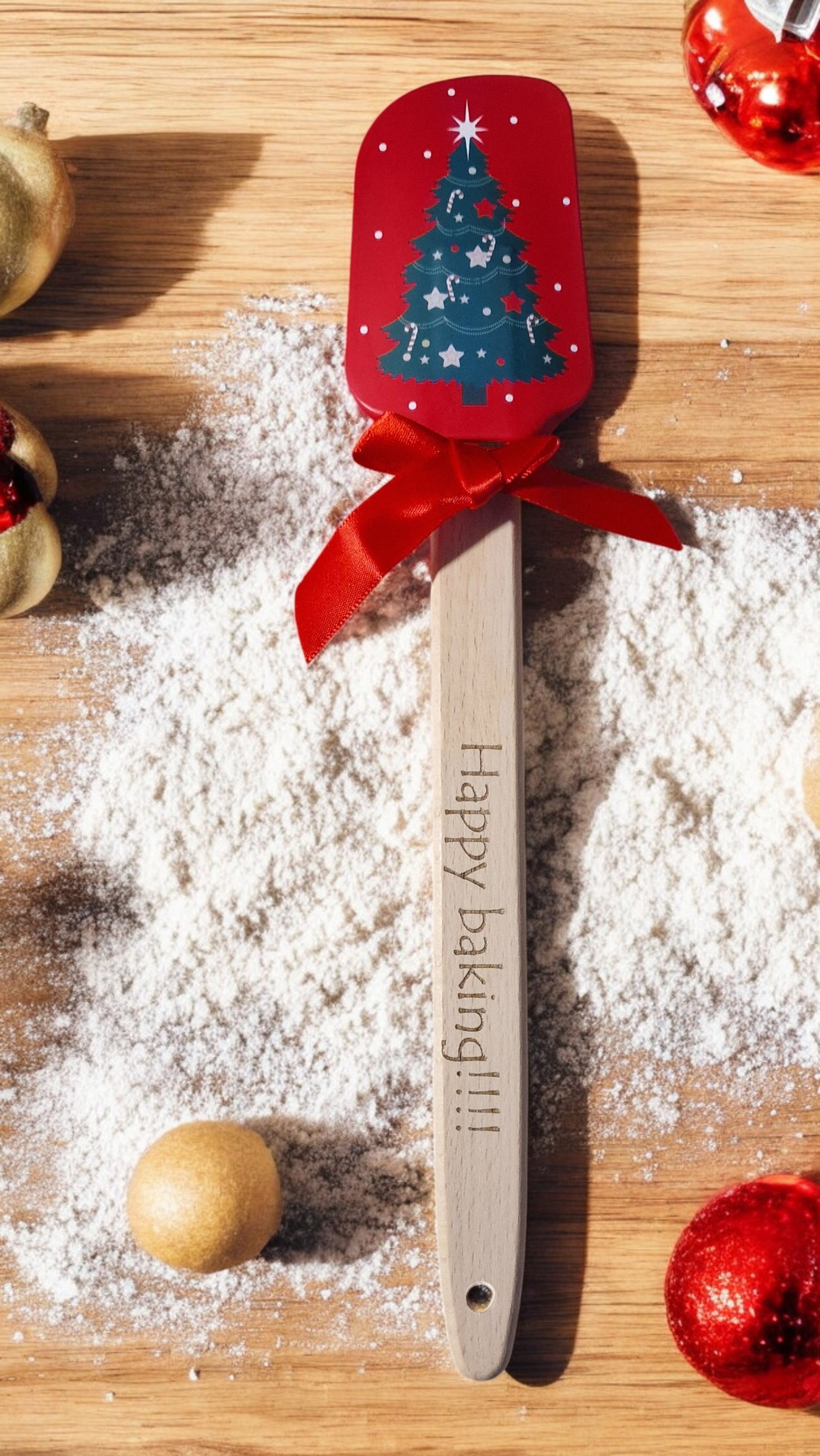 Spatula with Christmas design and 'Happy baking!!!' text on a wooden surface with flour and decorations.
