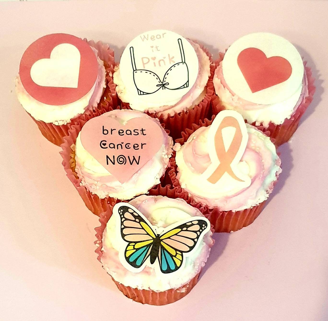 Assorted breast cancer awareness edible pre cut cake toppers featuring pink ribbons, hearts, butterflies, and supportive messages, arranged neatly on a light wood background.