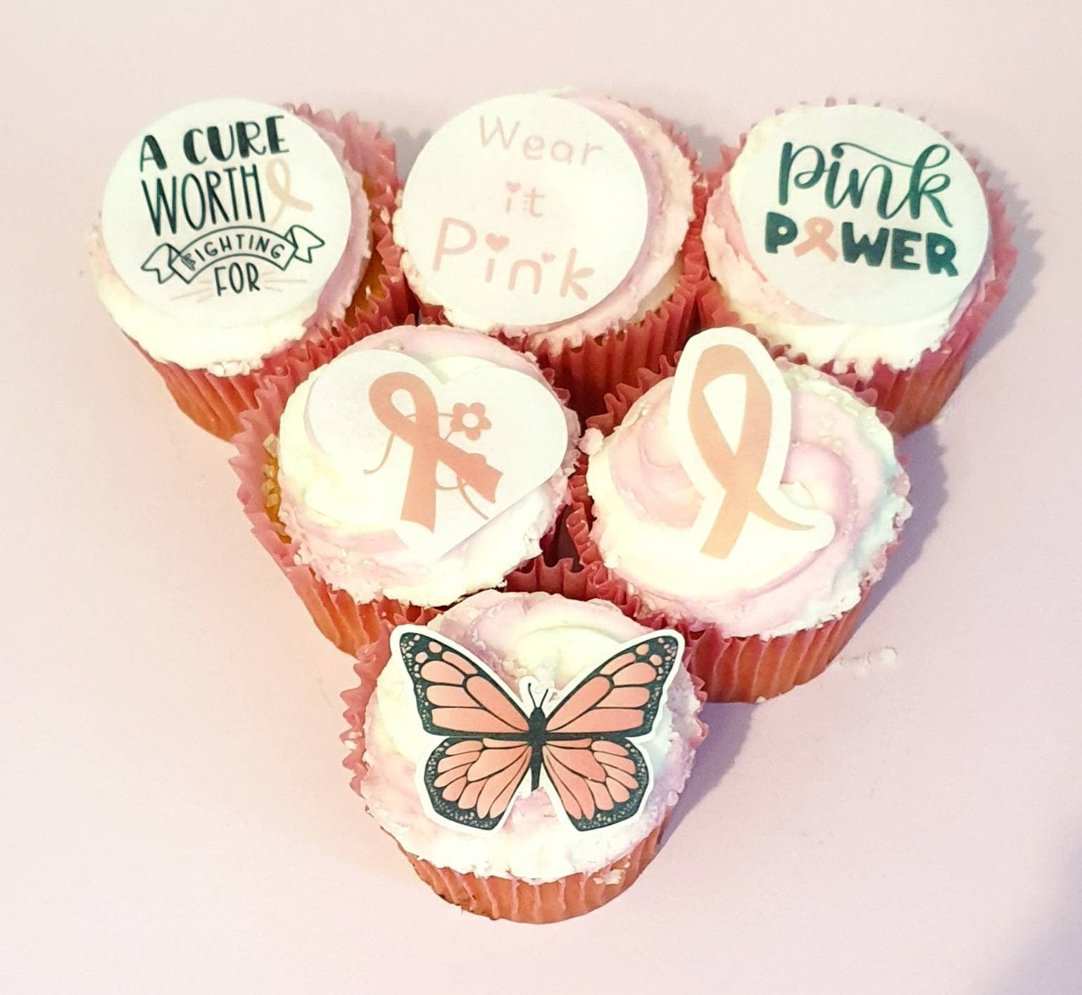 Assorted breast cancer awareness edible pre cut cake toppers featuring pink ribbons, hearts, butterflies, and supportive messages, arranged neatly on a light wood background.