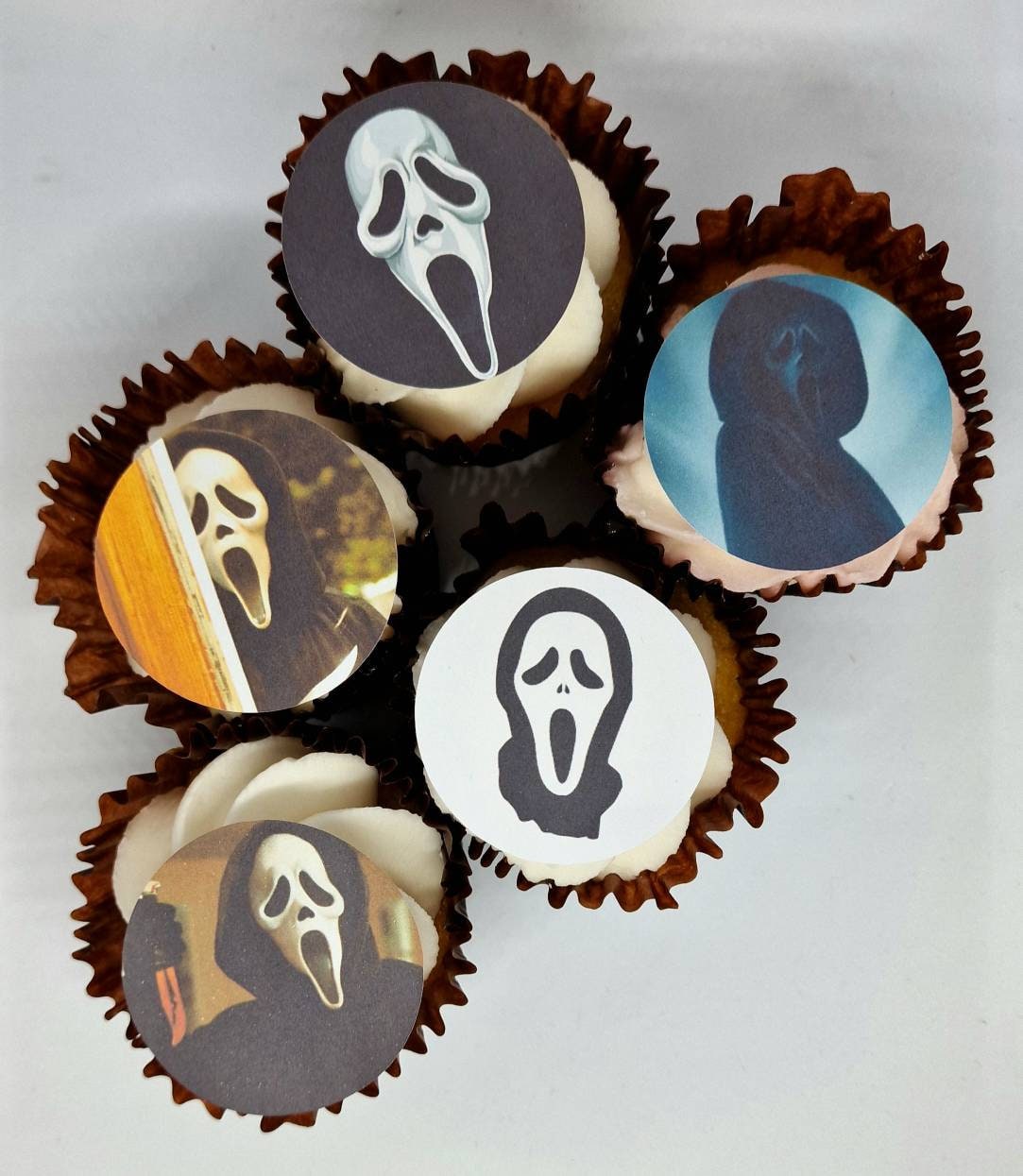Fifteen round edible precut cake toppers with Ghost Face mask designs and scenes inspired by the Scream movie, plus two knife-shaped edible toppers, all arranged on a light wooden background, each topper measuring 4.5cm