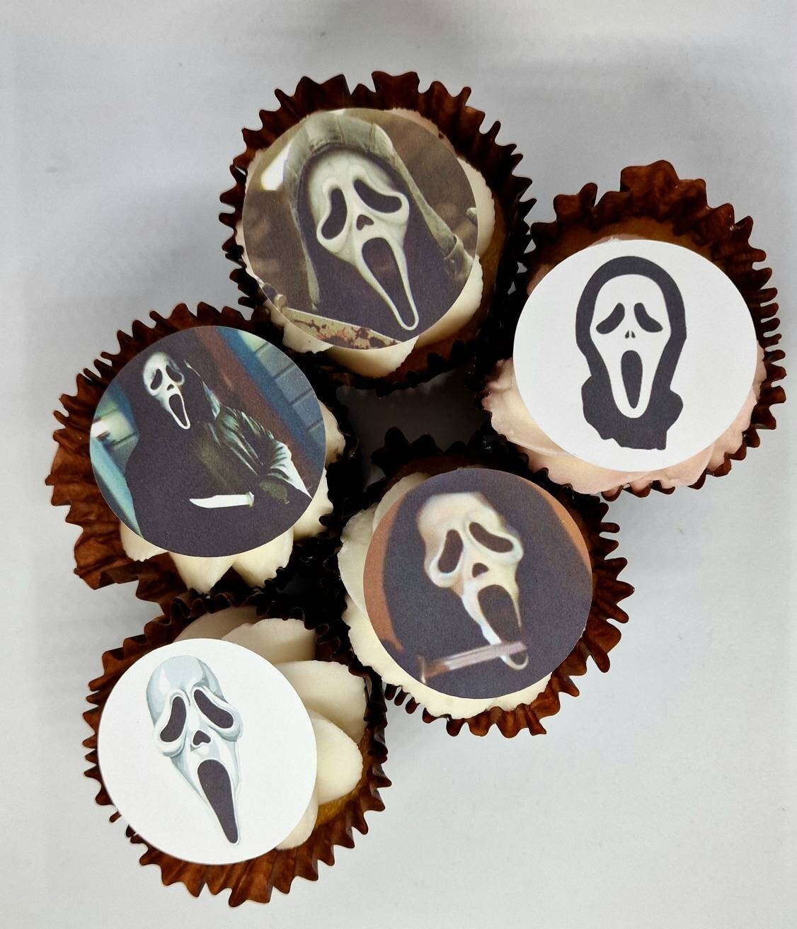 Fifteen round edible precut cake toppers with Ghost Face mask designs and scenes inspired by the Scream movie, plus two knife-shaped edible toppers, all arranged on a light wooden background, each topper measuring 4.5cm