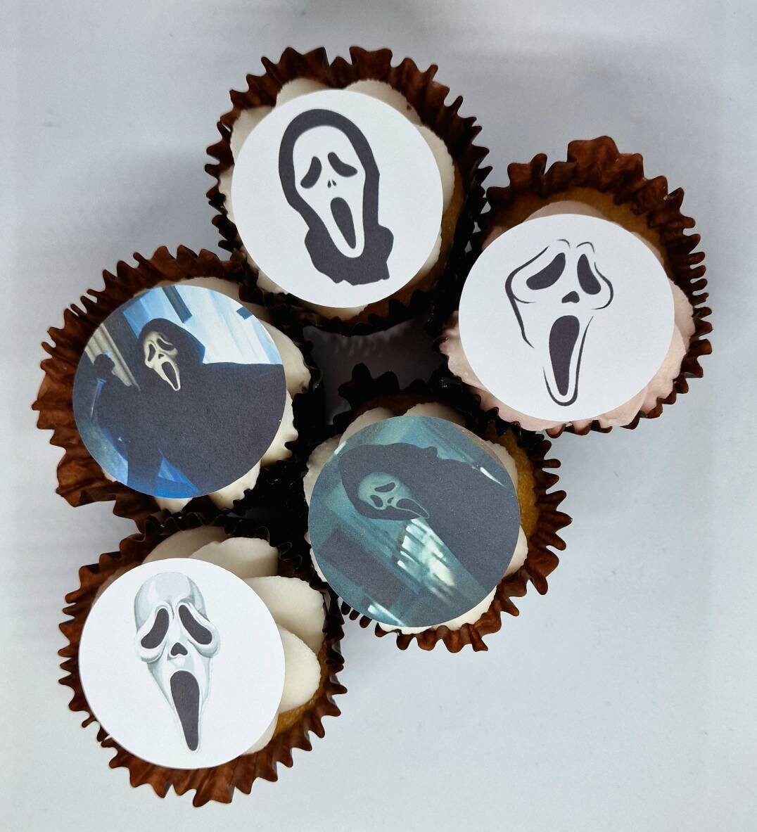 Fifteen round edible precut cake toppers with Ghost Face mask designs and scenes inspired by the Scream movie, plus two knife-shaped edible toppers, all arranged on a light wooden background, each topper measuring 4.5cm