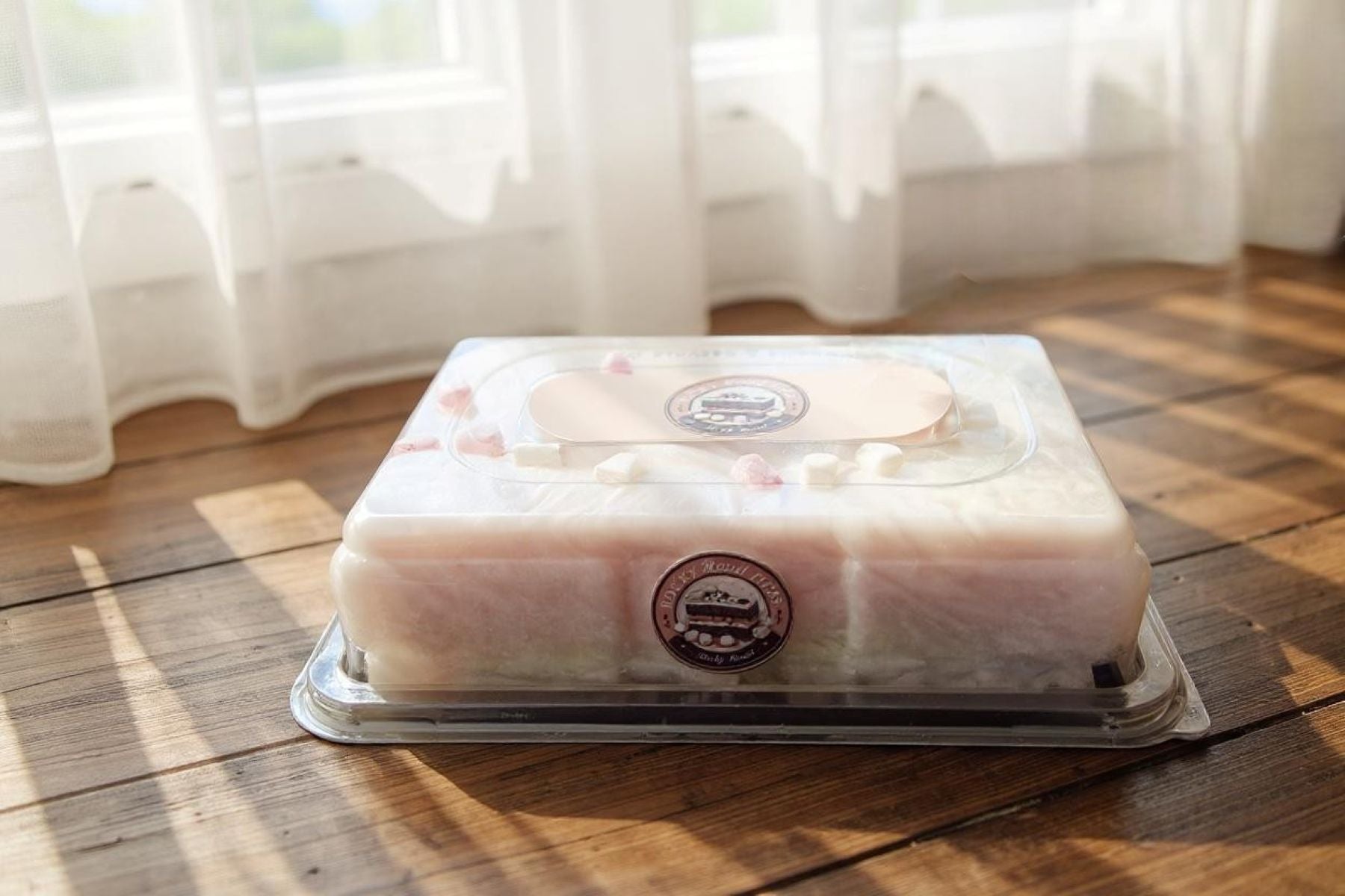 Rectangular cake in a clear plastic container on a wooden table with light curtains in the background