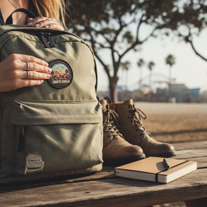 Green backpack with a patch, brown boots, and notebook on a wooden surface outdoors.