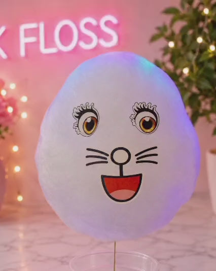 A white flossing mouth-shaped cotton candy holder on a clear stand, featuring a cartoon cat face with eyes, whiskers, and a red mouth. The product is displayed on a marble surface with a pink neon sign reading 'K FLOSS' in the background, flanked by potted plants with warm string lights.