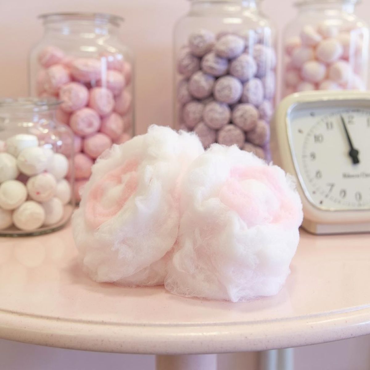 Decorative display with pink cotton candy, jars of pink and white spherical candies, and a clock on a white surface.