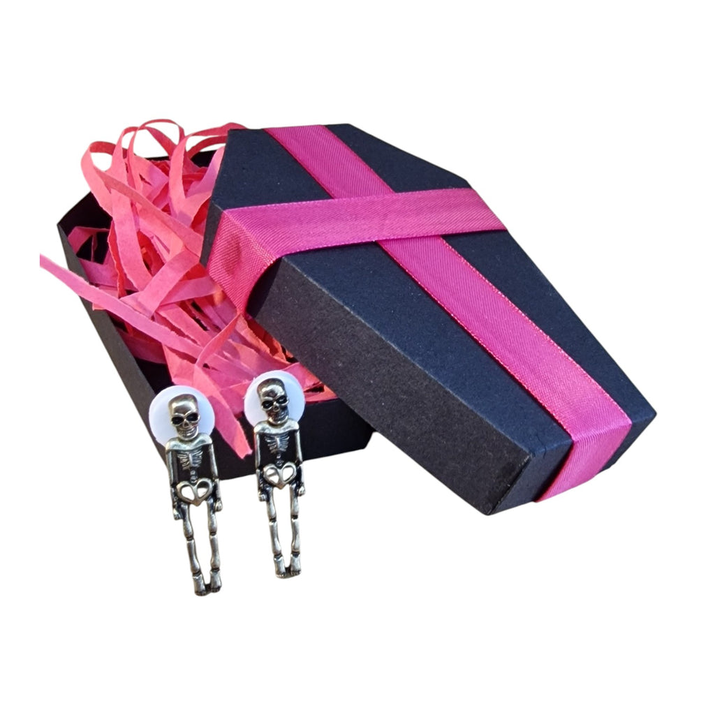 A pair of skeleton-shaped metal clips displayed in front of an open coffin-shaped gift box with bright pink ribbon and pink paper filler on a white background.