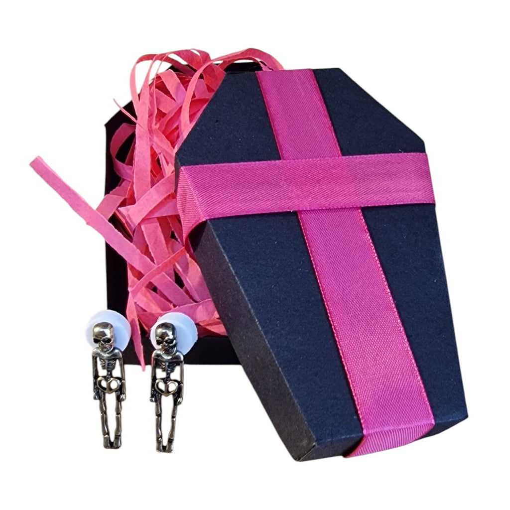 A coffin-shaped gift box in dark navy blue with a bright pink ribbon, filled with pink shredded paper, alongside two small skeleton clip-on earrings on a white background.