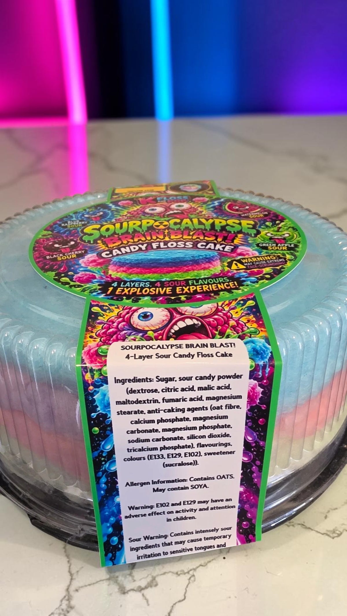 A round, multi-layered candy floss cake product labeled 'Sourpuss & Calypso Candy Floss Cake' with a colorful, psychedelic label and a clear plastic container holding pale blue layers, set on a marble surface with a pink ambient light in the background.