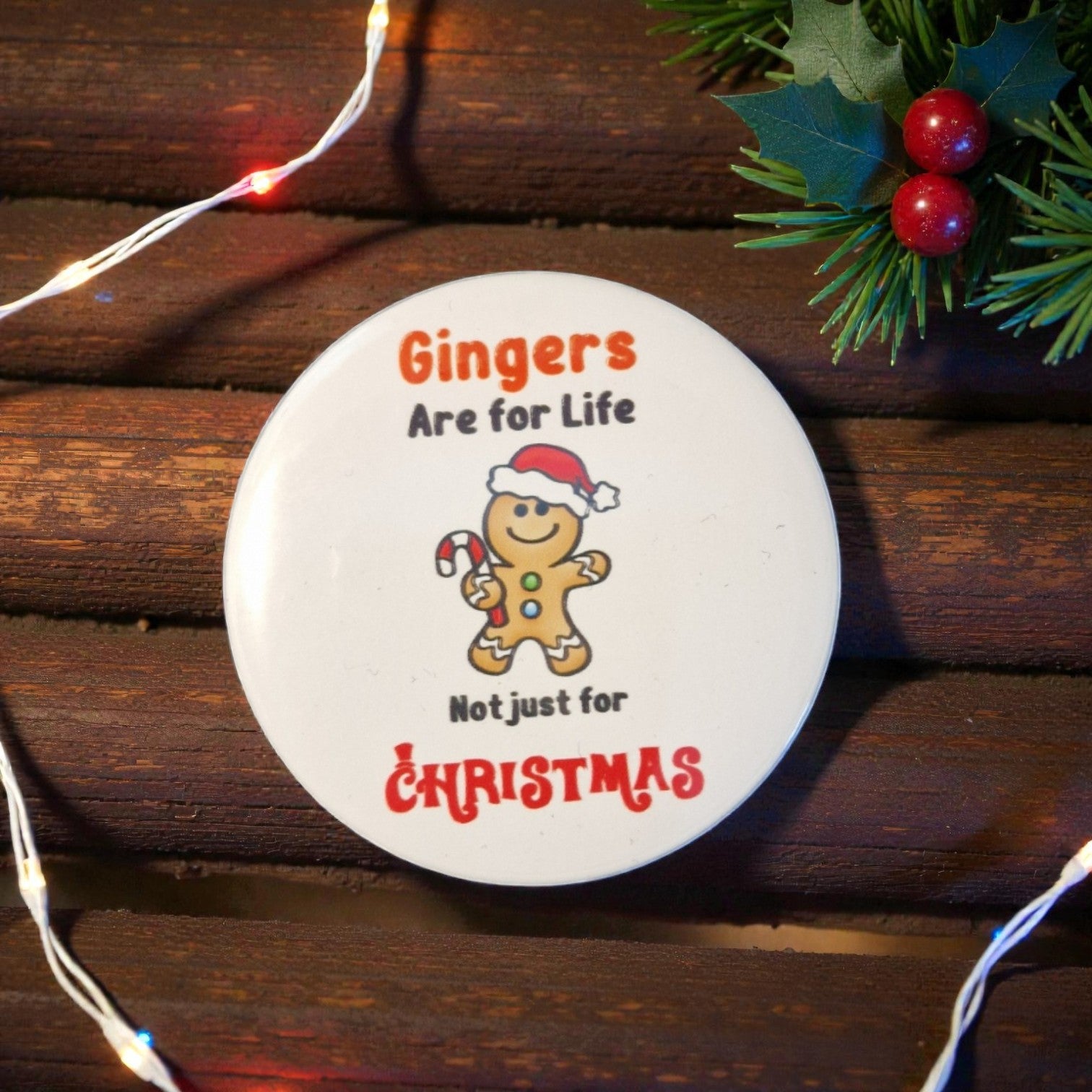 Round Christmas ornament with gingerbread man and text on a wooden surface with holly and lights.