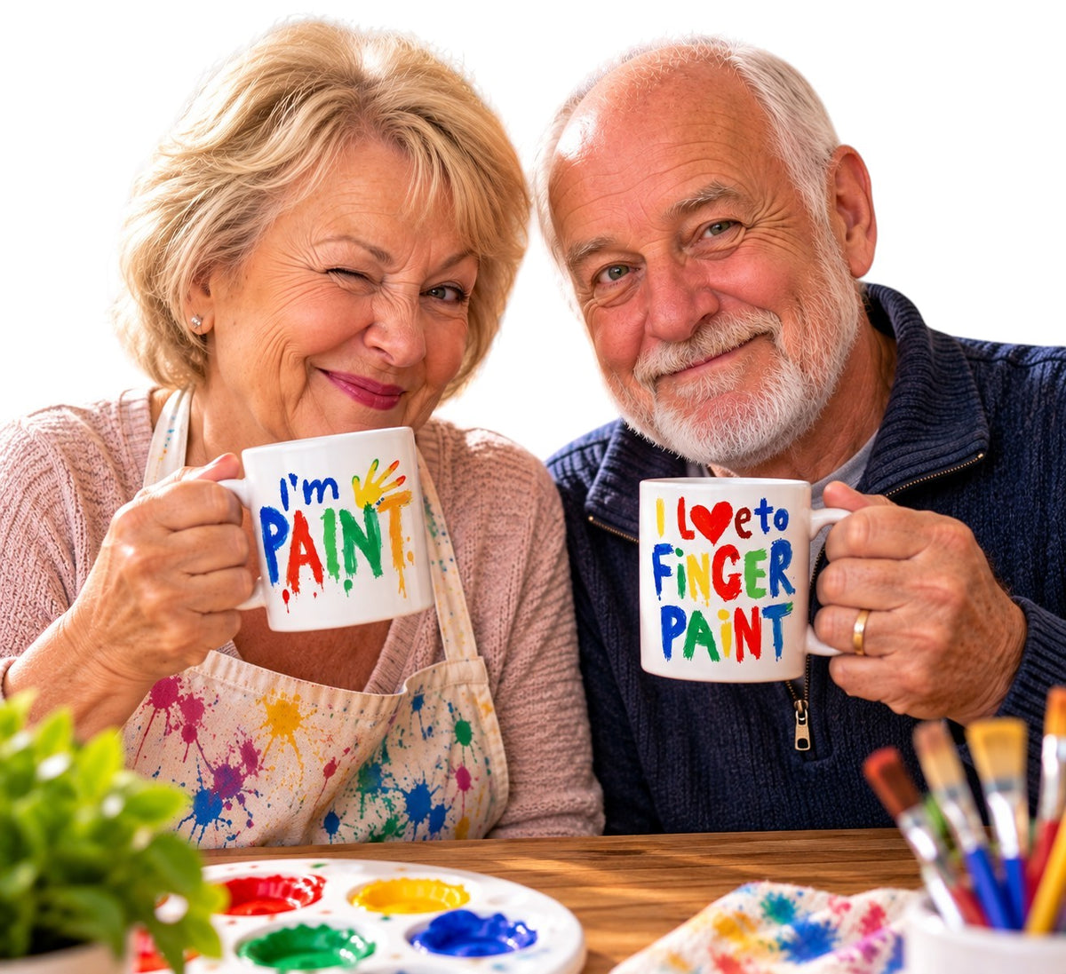 An older couple smiling while holding white mugs that say 'I'm PAINT' and 'I ♥ to FINGER PAINT' in colorful lettering, with paint splatters on their aprons and paintbrushes on a table in front of them.