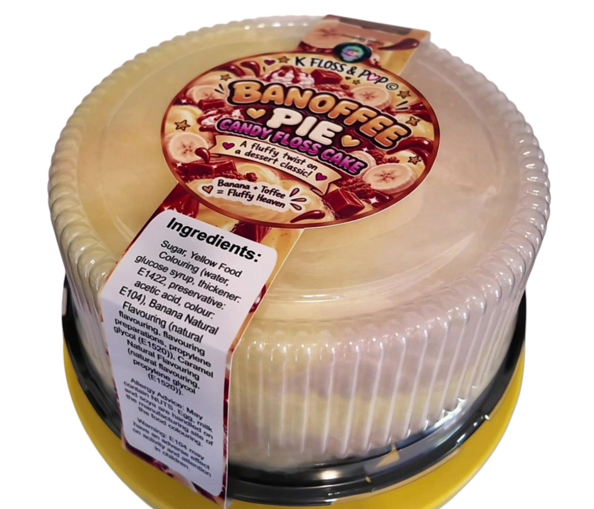 A round plastic tub of Kloss & Pypa's Banoffee Pie Candy Floss Cake, showing the creamy yellow cake inside, a clear lid with a colorful red and brown label, and a white ingredient tag on a yellow base.