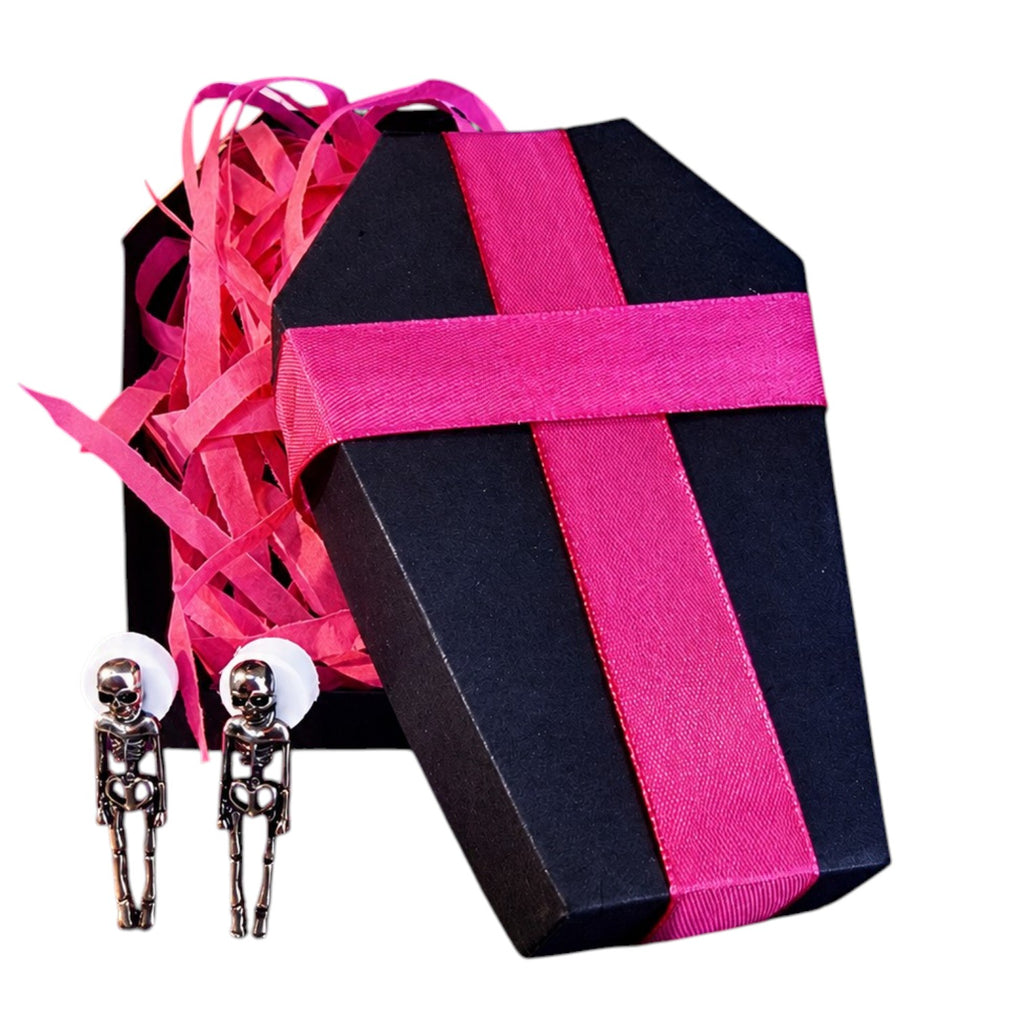A coffin-shaped black gift box with a bright pink ribbon and pink shredded paper filler, shown alongside a pair of silver skeleton stud earrings on a white background.