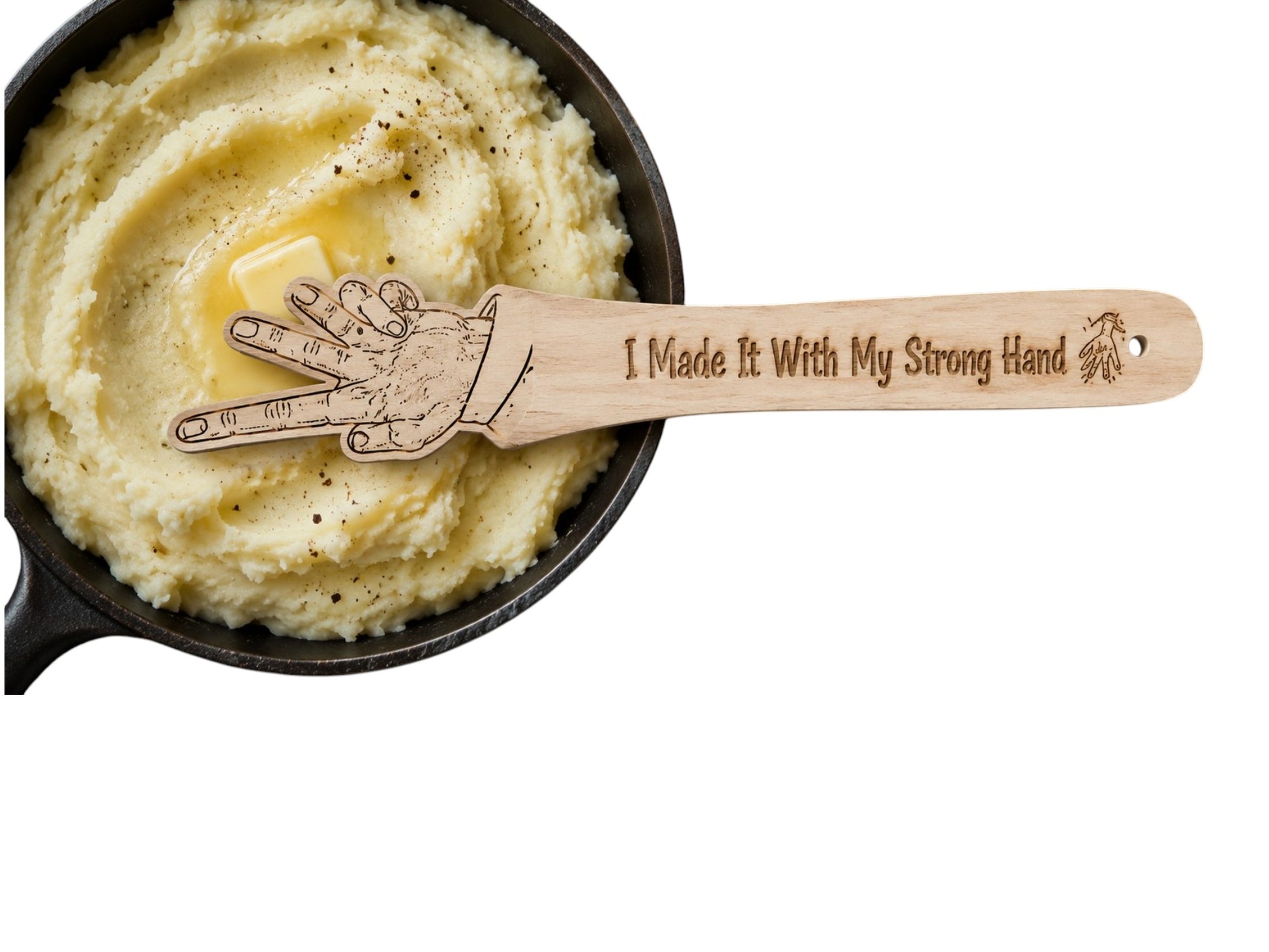 A wooden spatula with a hand graphic and the engraved text 'I Made It With My Strong Hand' resting on top of mashed potatoes in a black skillet with a pat of butter, isolated on a white background.