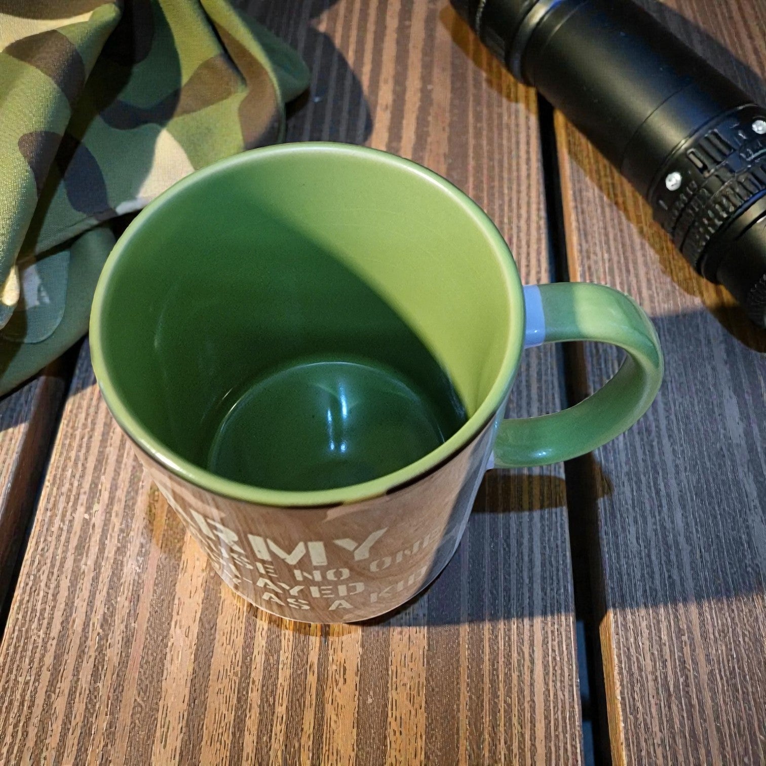 Green mug on a wooden surface with a camouflage-patterned bag and a camera in the background.