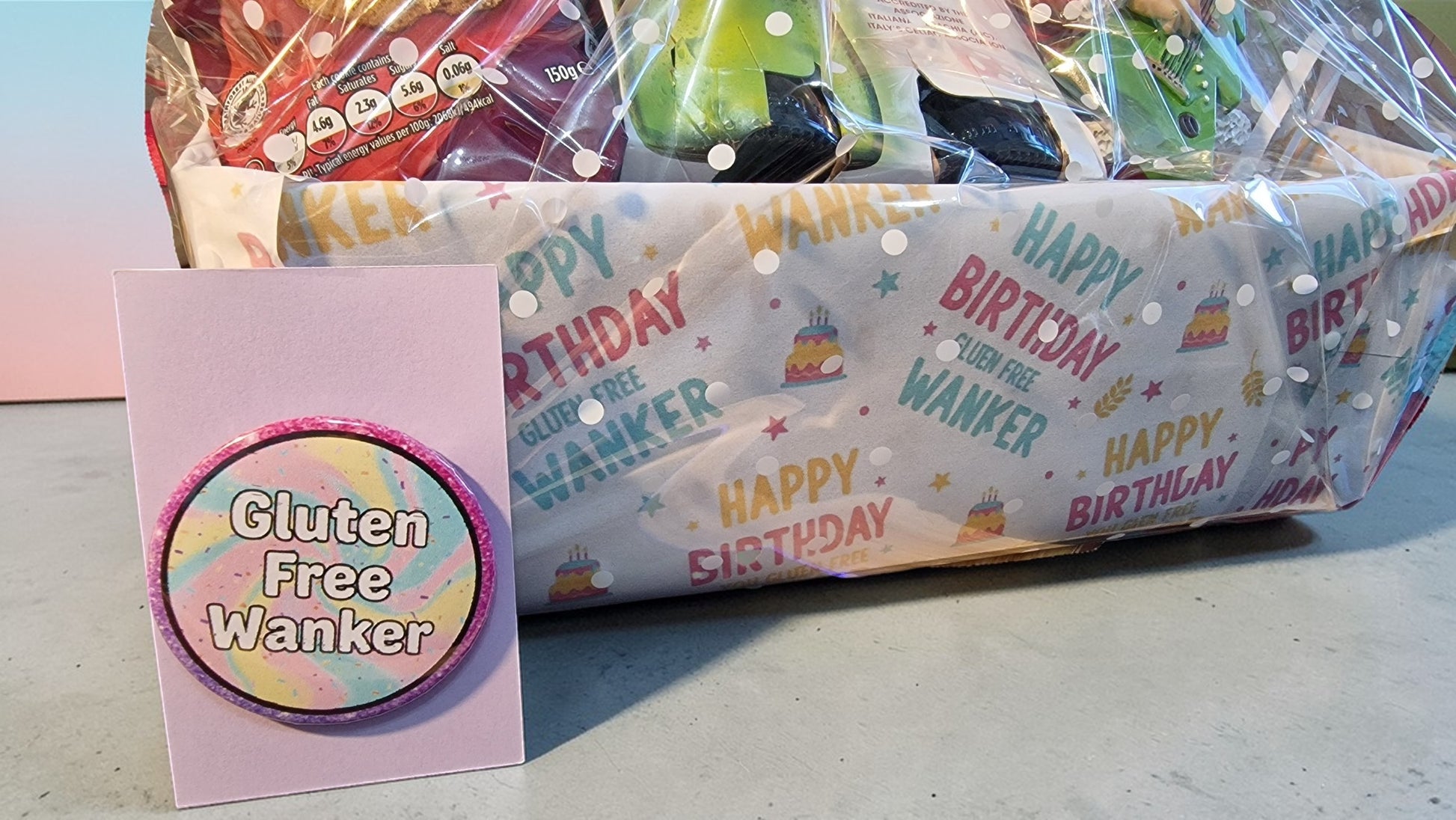 Gift basket with 'Happy Birthday' wrapping and a card labeled 'Gluten Free Wanker' on a concrete surface.