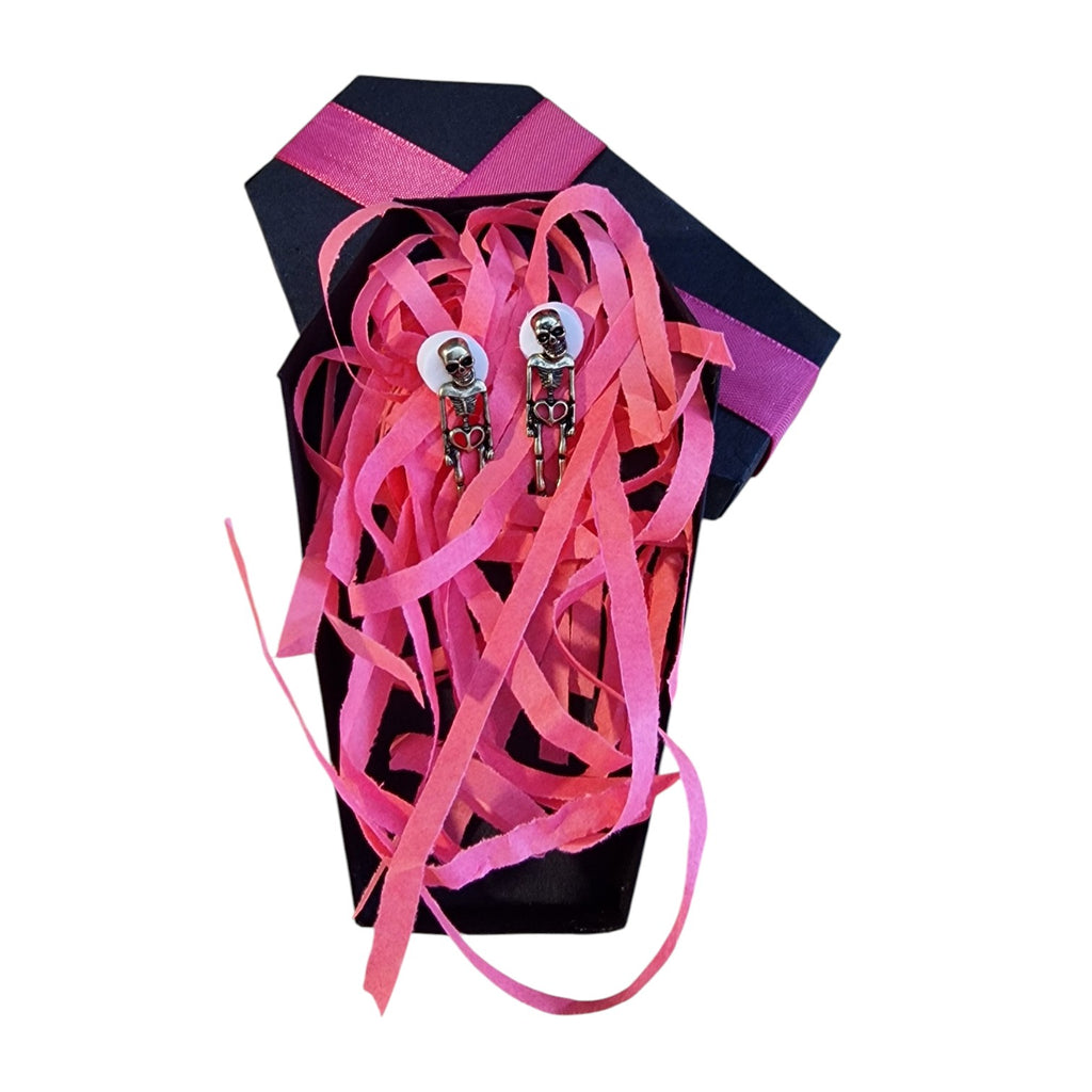 A pair of gothic skeleton stud earrings displayed inside a black coffin-shaped gift box filled with pink and magenta shredded ribbon, set against a plain white background.