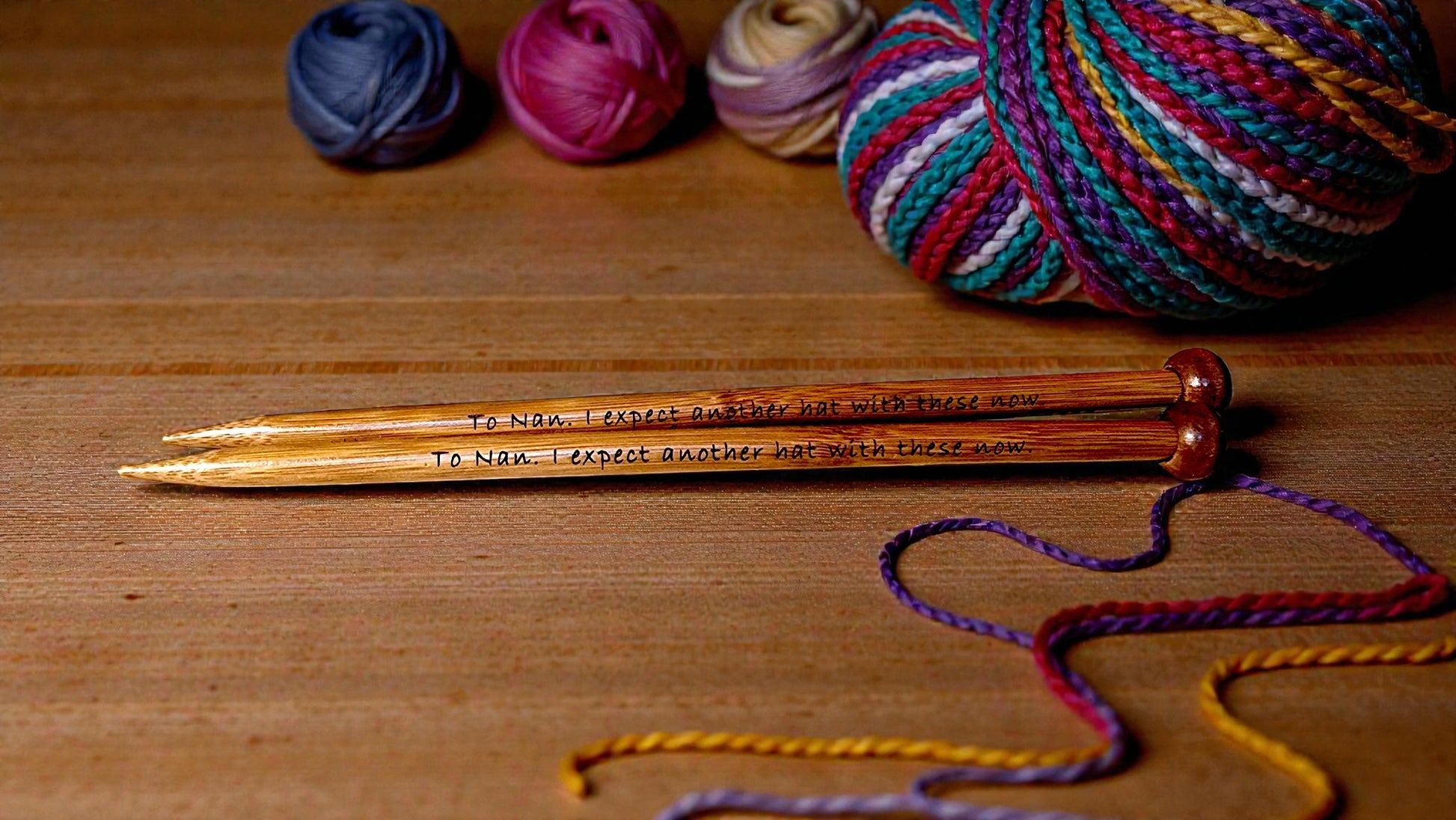 Pair of personalised bamboo knitting needles with engraved messages, chunky wool wrapped around, displayed on a wooden table with colourful yarn in the background