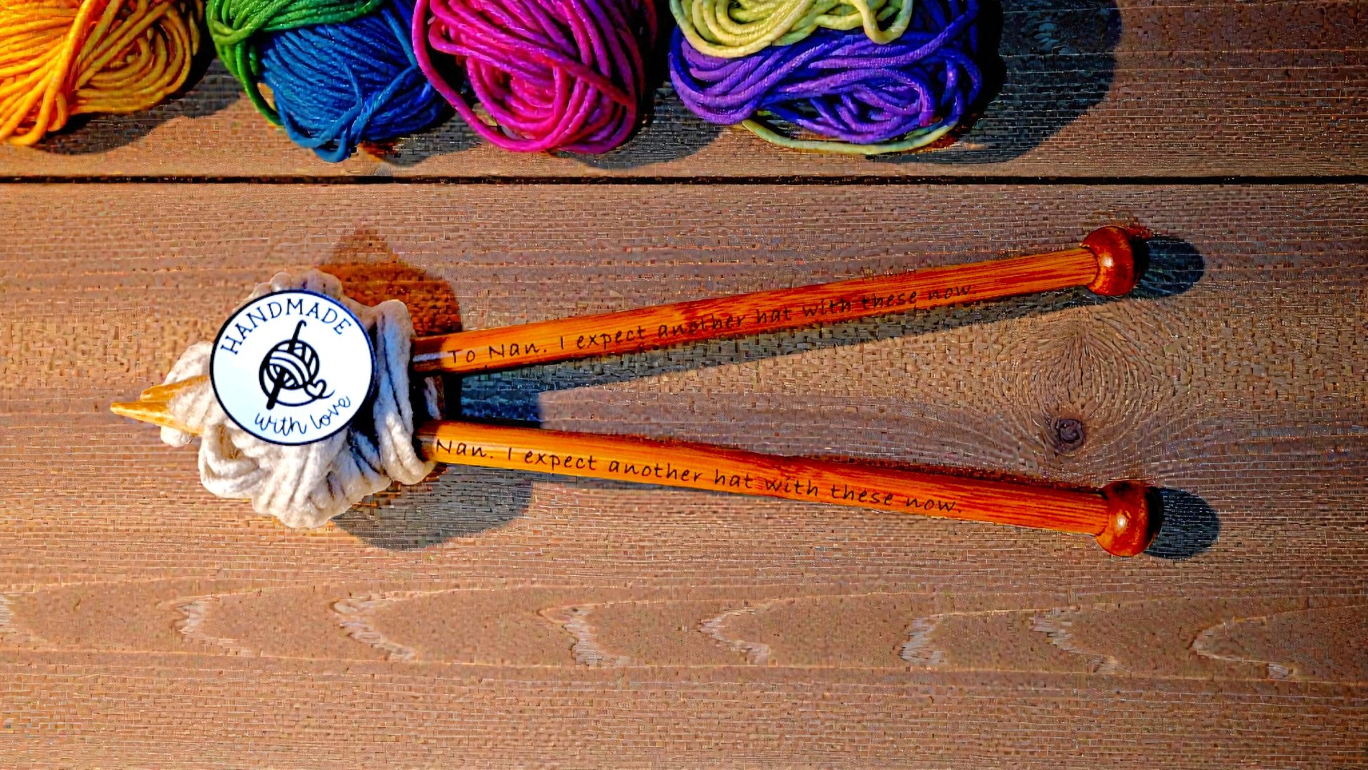 Pair of personalised bamboo knitting needles with engraved messages, chunky wool wrapped around, displayed on a wooden table with colourful yarn in the background