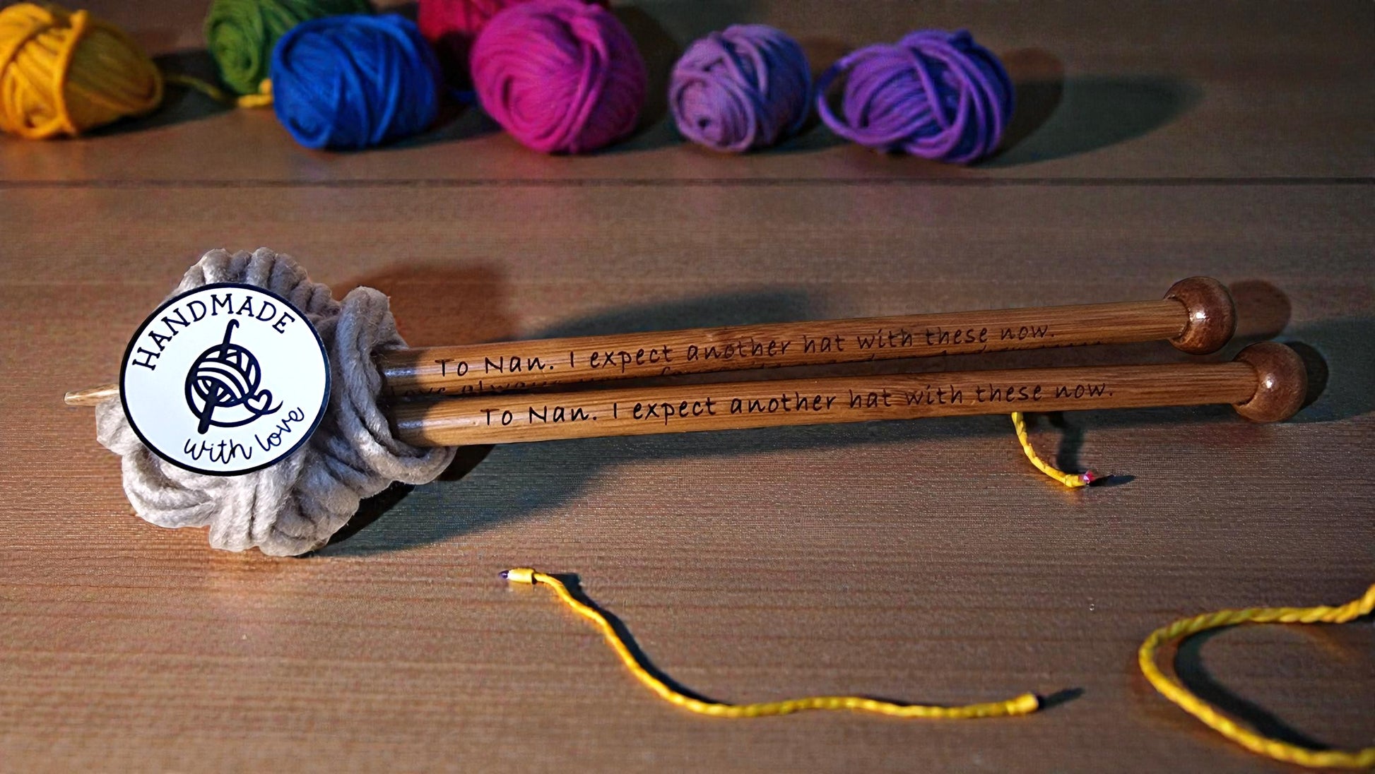 Pair of personalised bamboo knitting needles with engraved messages, chunky wool wrapped around, displayed on a wooden table with colourful yarn in the background