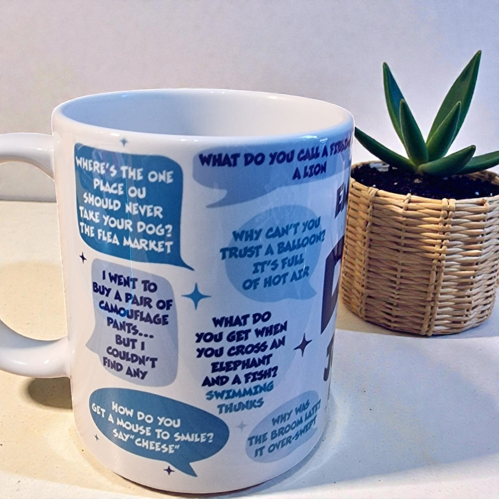 White mug with blue text and graphics on a light surface with a plant in the background