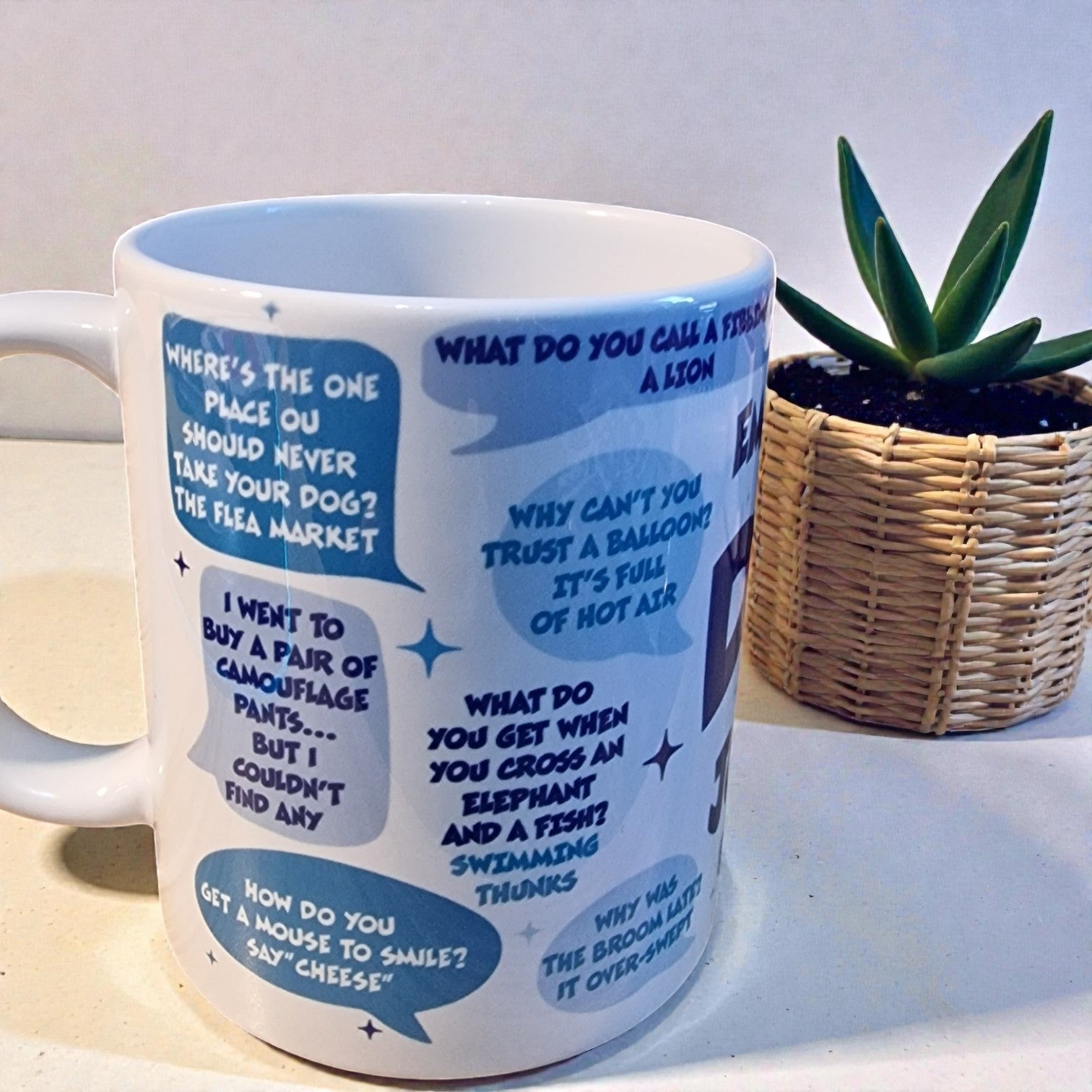 White mug with blue text and graphics on a light surface with a plant in the background