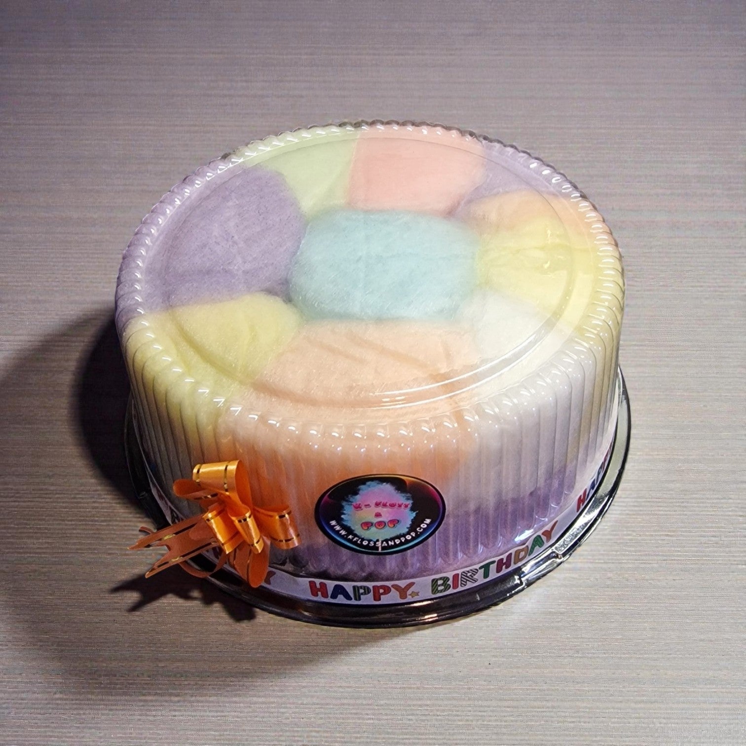 Pastel-colored cake in a clear plastic container with a 'Happy Birthday' label on a gray surface.