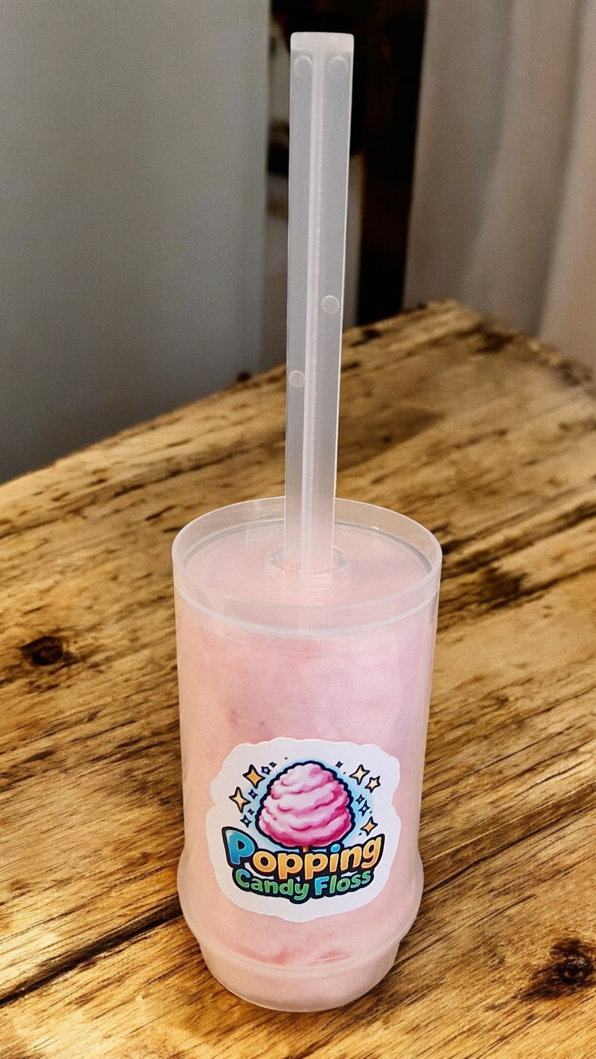 Pink drink in a cup with a straw on a wooden surface, featuring a 'Popping Candy Floss' label.